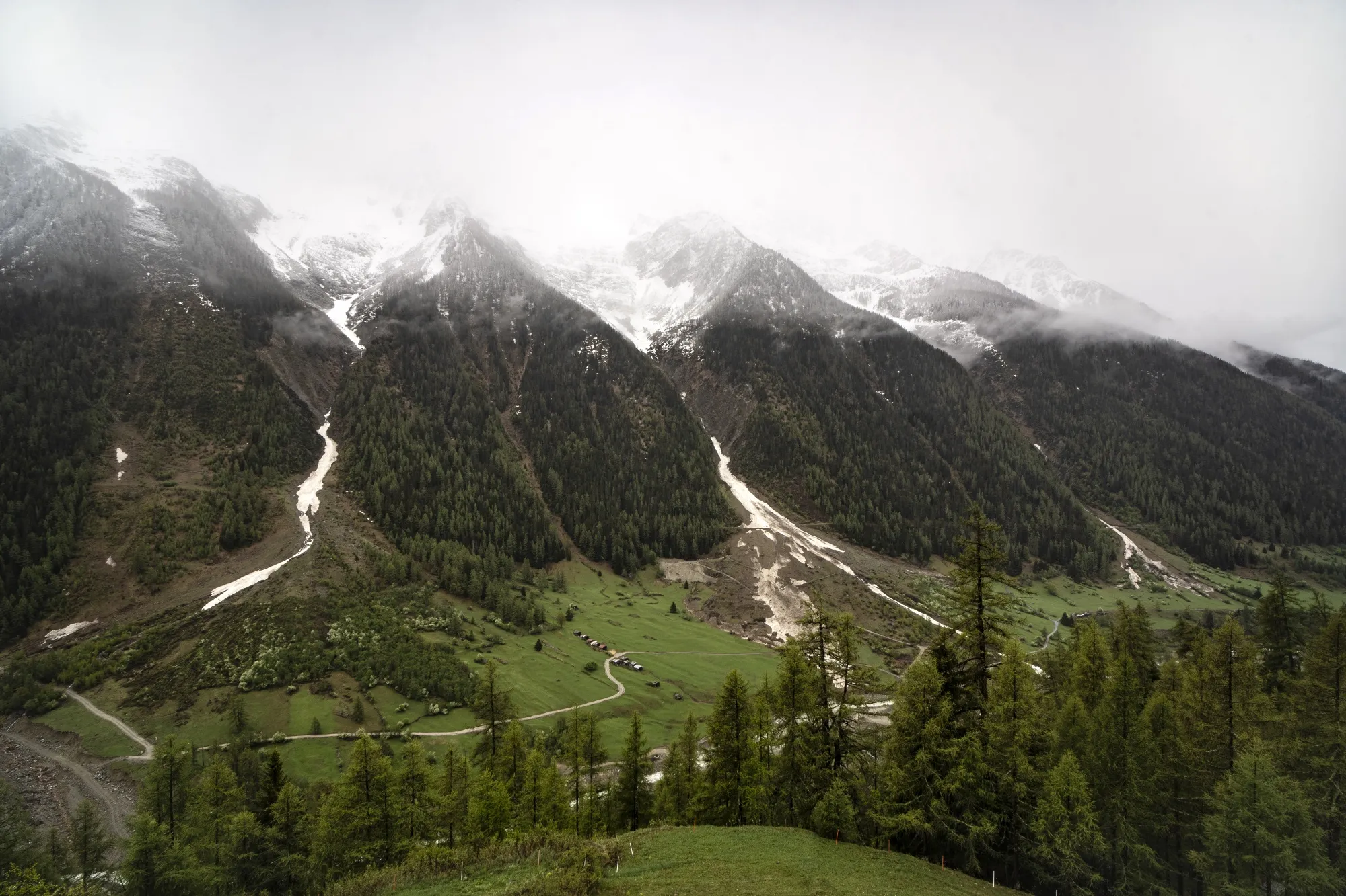 Tall mountains, topped with snow, disappear into clouds. A few buildings dot the grassy slopes below.