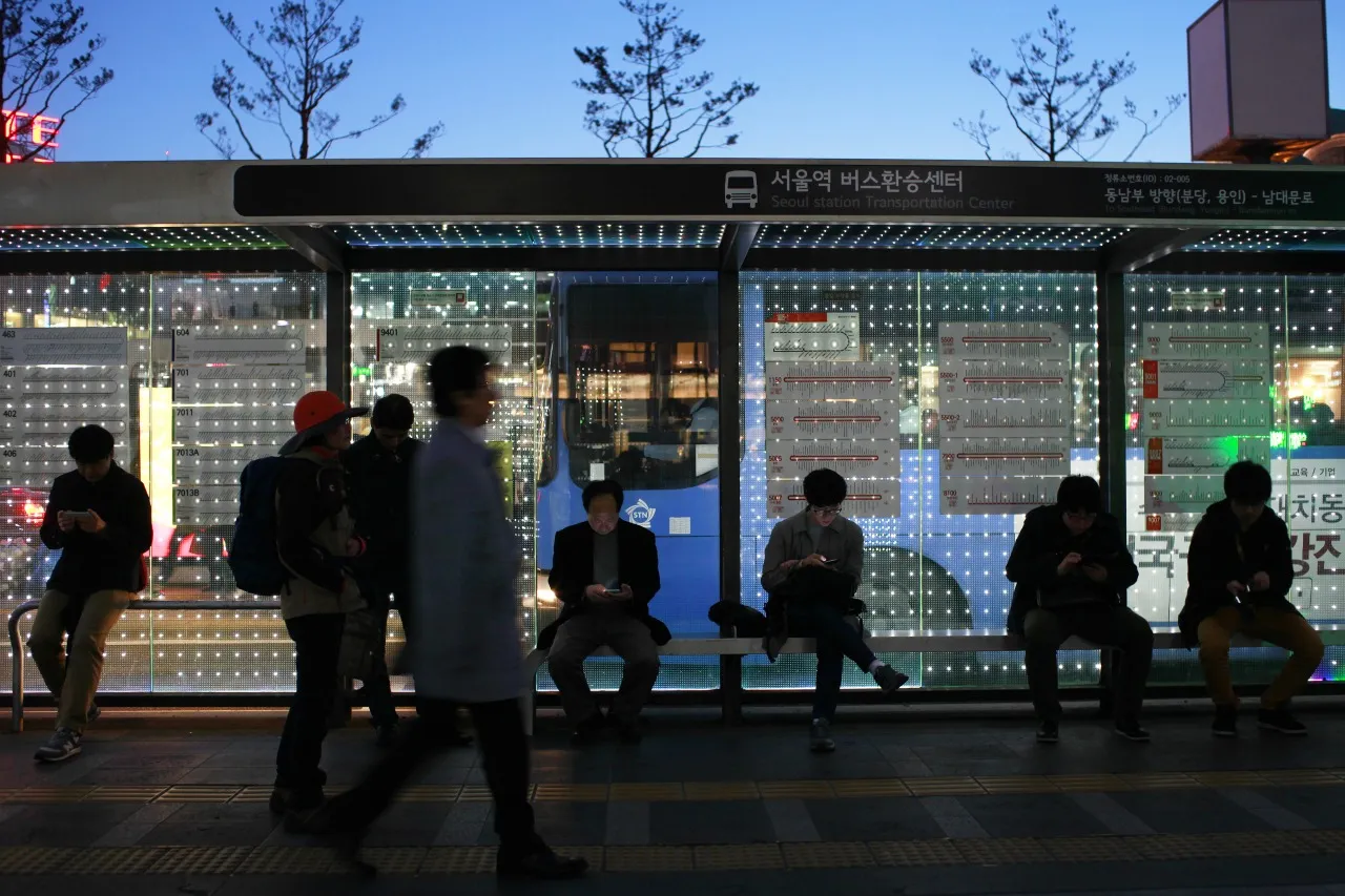 Commuters use mobile devices at a bus stop near Seoul Station at dusk in Seoul, South Korea.