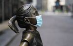 Fearless Girl, with a PPE mask on in front of the New York Stock Exchange in the Wall Street Financial District of Manhattan in NY.