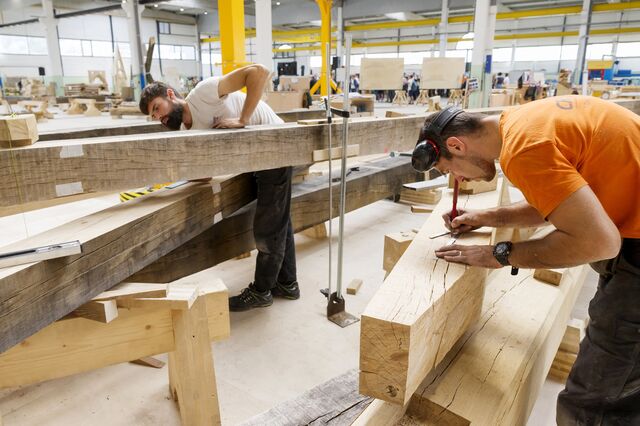 Carpenters in a brightly lit workshop work on wooden beams