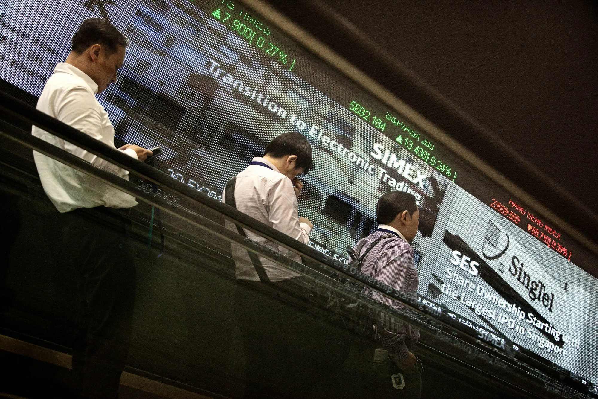 Men ride on an escalator past an electronic screen and ticker board displaying stock figures at the Singapore Exchange Ltd. (SGX) headquarters in Singapore, on Thursday, Jan. 19, 2017. The SGX has tendered a 4.75% stake in the Bombay Stock Exchange for sale in an initial public offer, valuing its shares at S$42.8m to S$42.9m, the exchange said in a filing today.