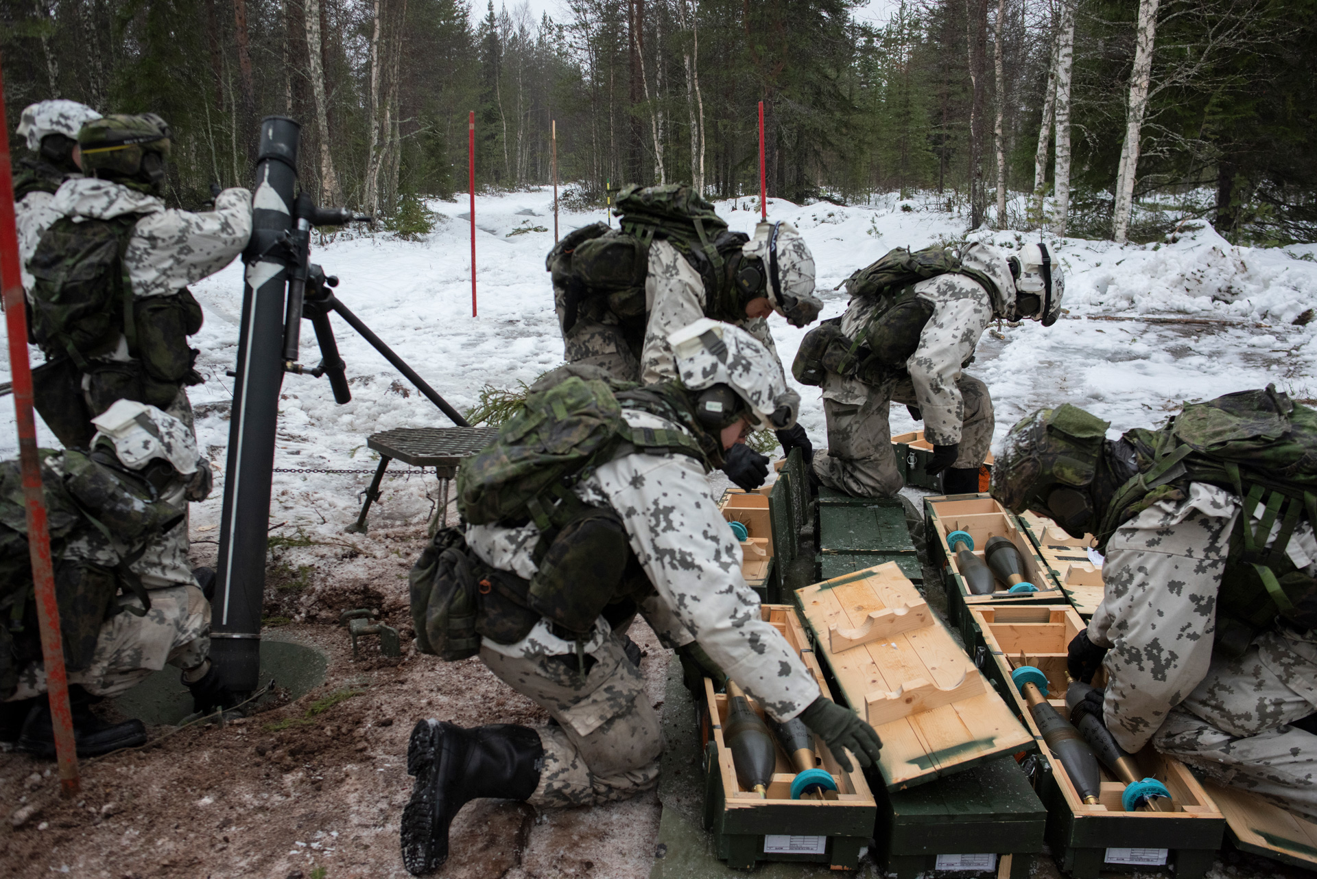 A platoon of seven Finnish soldiers, all wearing white camouflage uniforms, are manning a mortar position in a forest. Three are around the mortar tube, which rests pointing upward on a tripod and is as tall as the soldier standing next to it. The others are off to one side, unpacking mortar shells from wooden crate s. The ground is covered with snow.