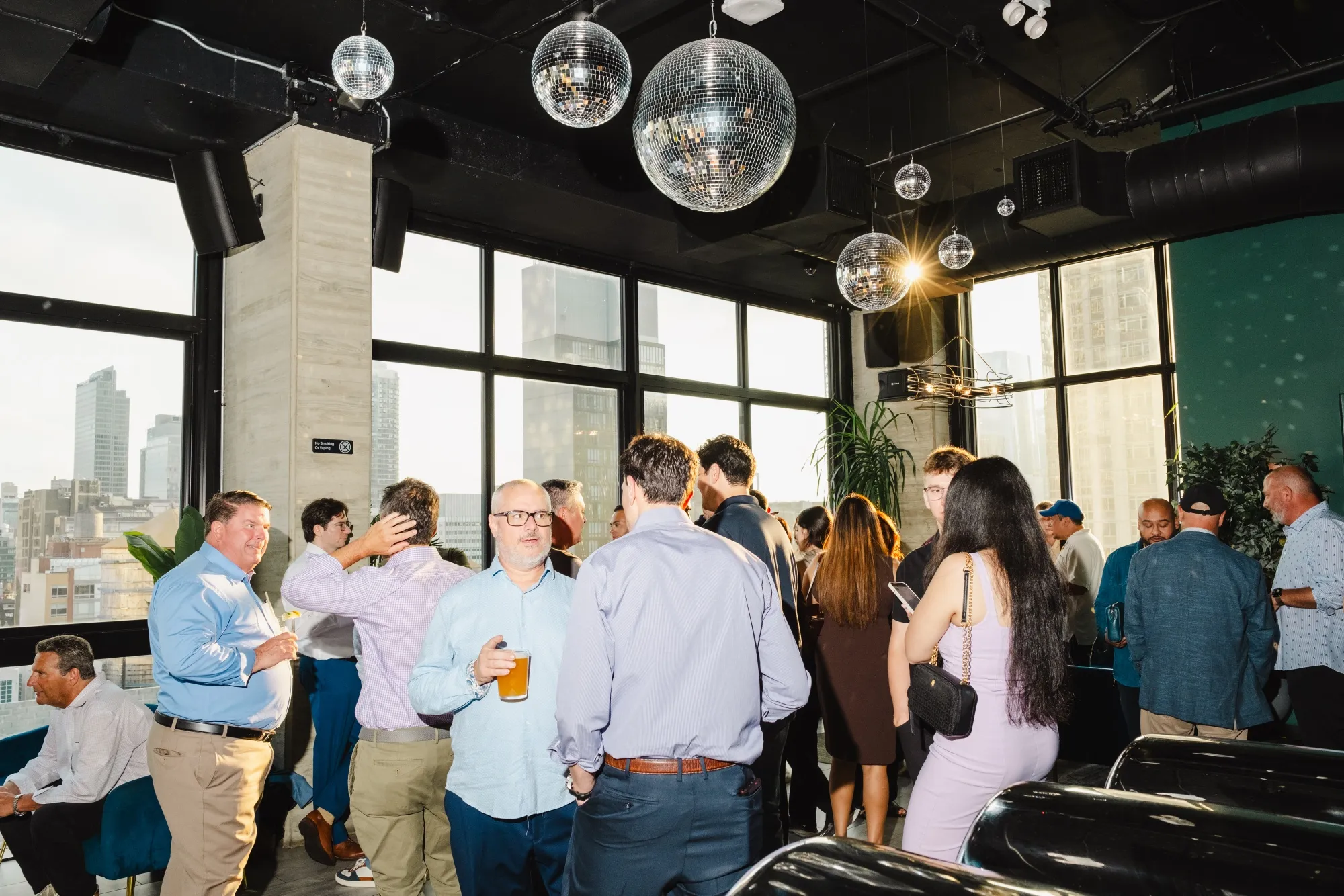 Several people chat in a room with disco balls overhead and large windows that show New York City skyscrapers in the background.
