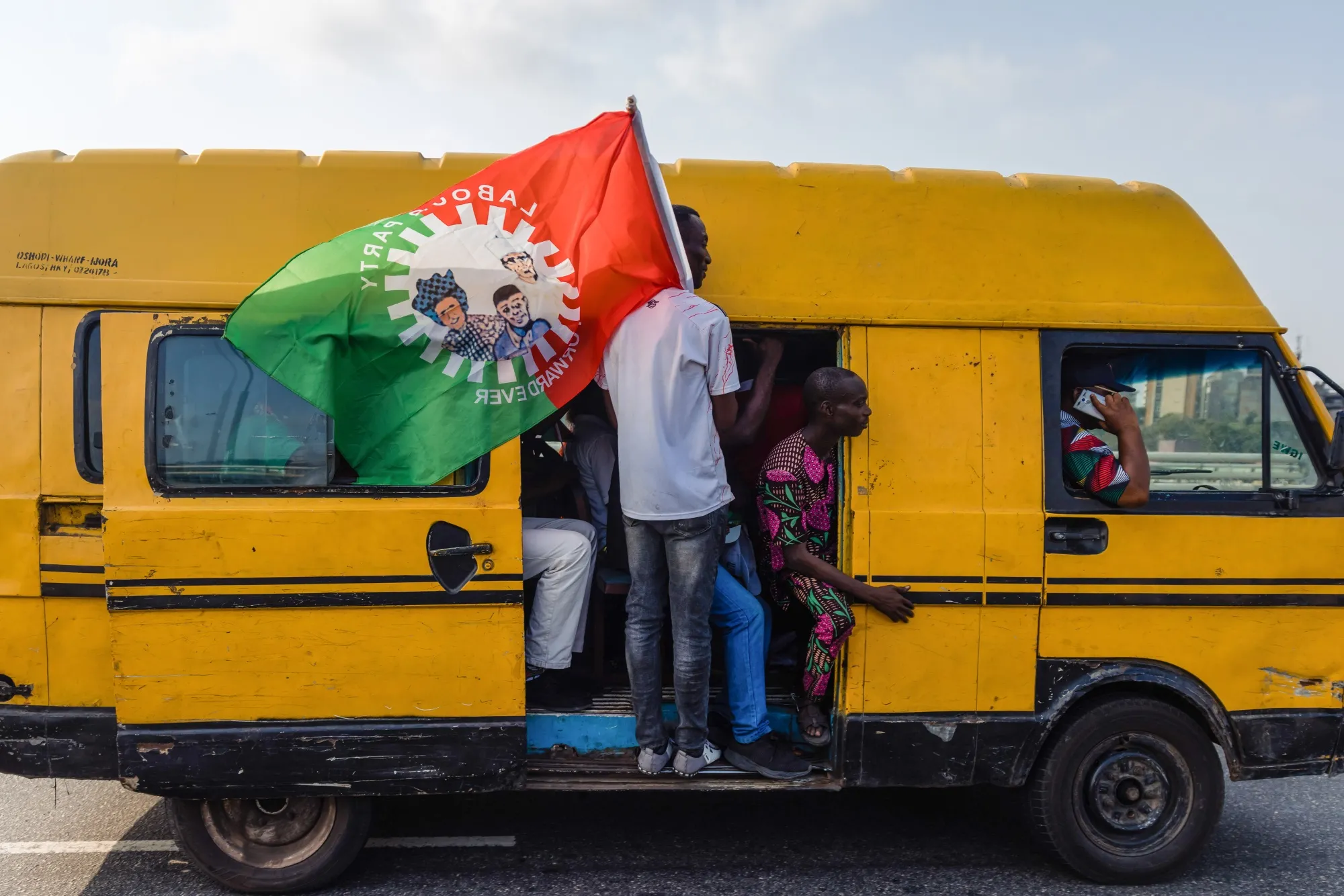 A supporter rides on the side of a minibus holding a Labour Party flag.