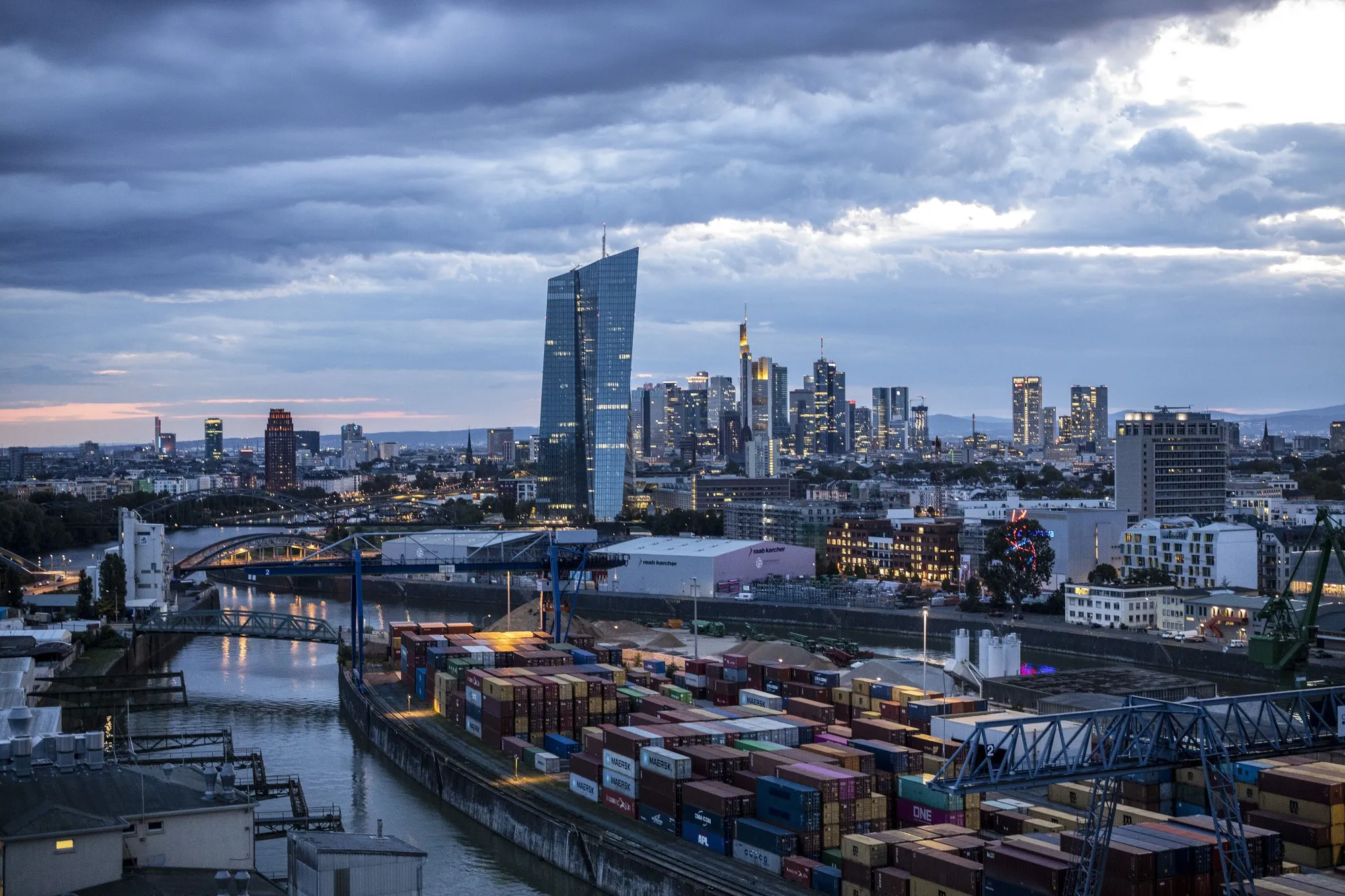The European Central Bank (ECB) headquarters, center, stand illuminated back dropped by the financial district skyline beyond shipping containers on the quayside at Frankfurt Osthafen container dock, operated by Contargo Rhein-Neckar GmbH, in Frankfurt, Germany, on Tuesday, Aug. 13. 2019. Germany’s economy shrank in the second quarter, ramping up pressure on Chancellor Angela Merkel to unleash fiscal stimulus as manufacturers reel from a U.S.-China trade war.