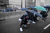 Multiple protesters wearing helmets and gas masks crouch down in the street holding umbrellas overhead for protection from possible falling objects