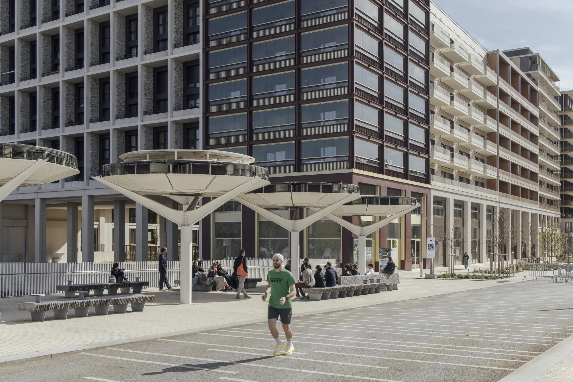 A photo shows a modern urban development featuring a mix of contemporary architectural styles and active public space. In the background, a series of tall buildings display diverse facades, ranging from a white grid-like structure to a darker building with large glass windows and another with long, repetitive balconies. The foreground is dominated by a wide, sunlit pedestrian area with striped pavement where a man in a green t-shirt is jogging toward the camera. Several people are resting on long concrete benches beneath a row of unique, futuristic white sunshades shaped like wide, flat-topped funnels.
