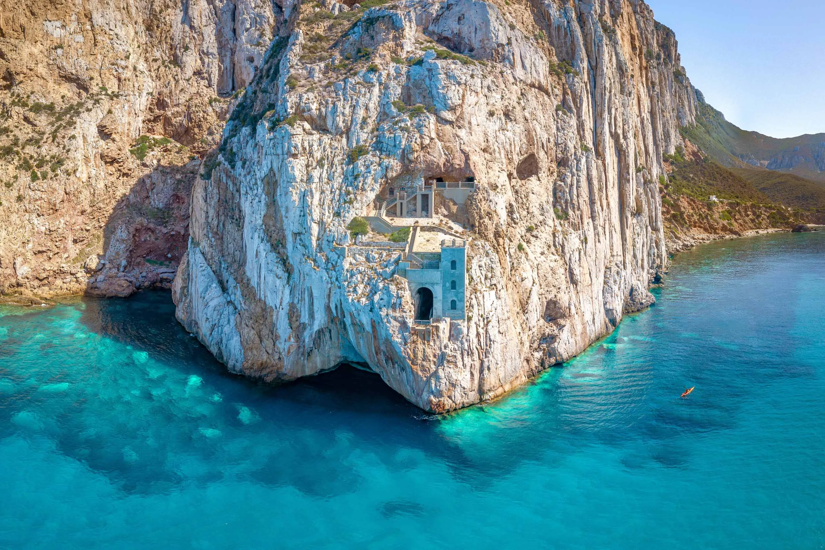 A solitary window and small structure carved into the middle of large white and green cliffs above the crystal waters of the Sea of Sardinia.