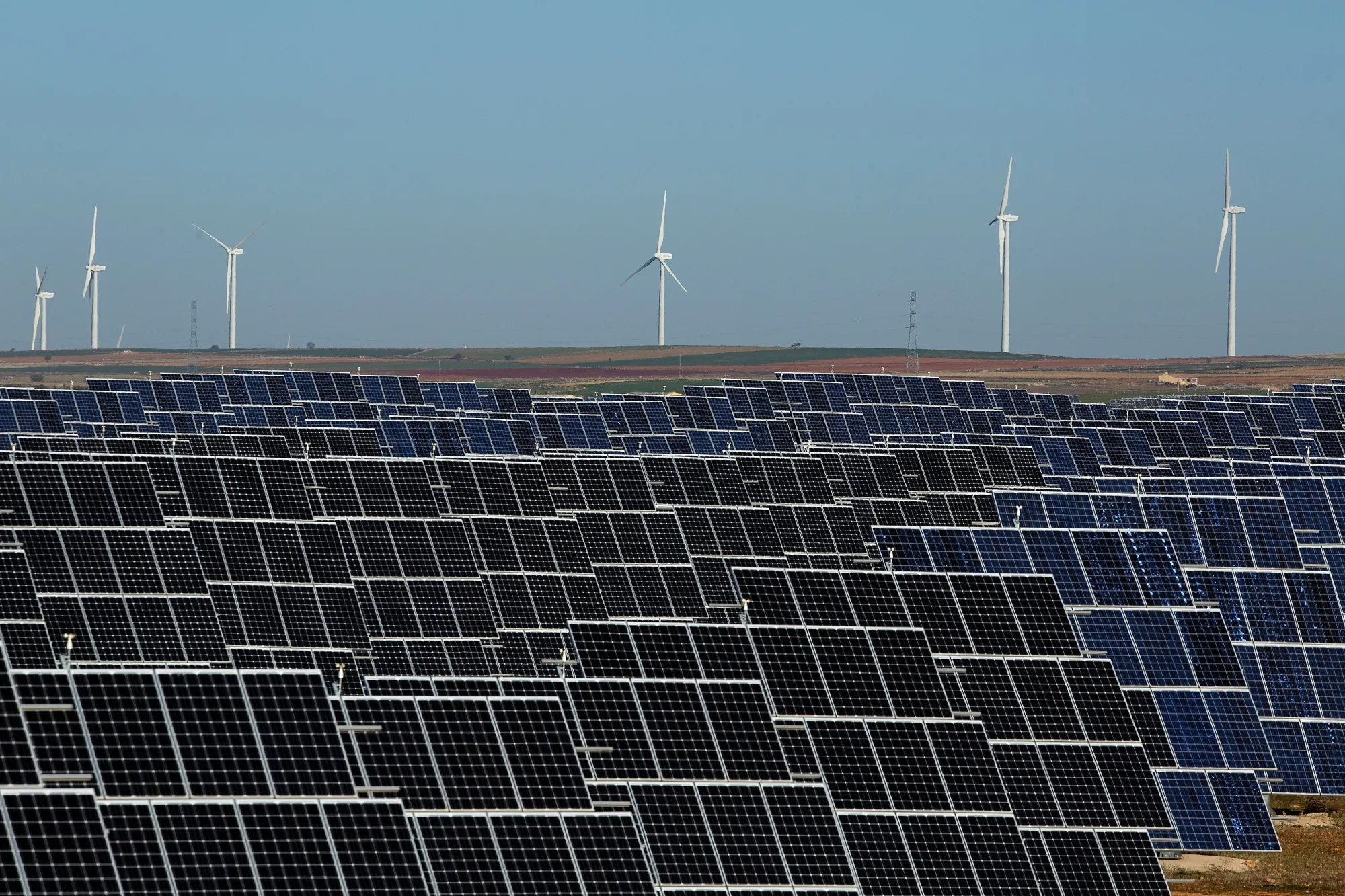 &nbsp;power panels stand at Abaste's El Bonillo Solar Plant while wind turbines spin at a wind farm on the background.