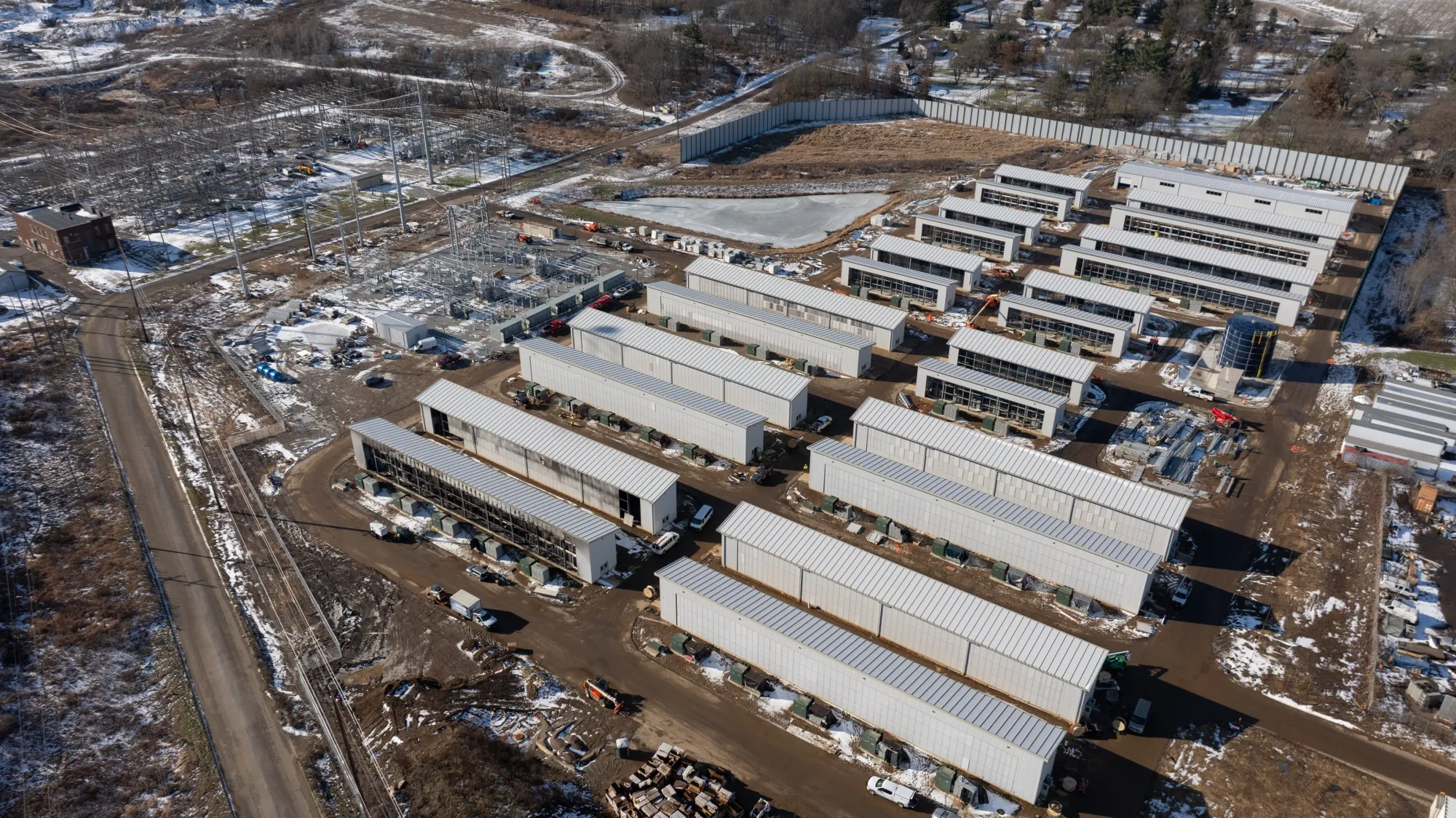 An aerial shot of rows of long narrow structures. One such structure in the front appears to show fire damage. Some snow mixes with the mostly dirt ground surrounding the construction site.