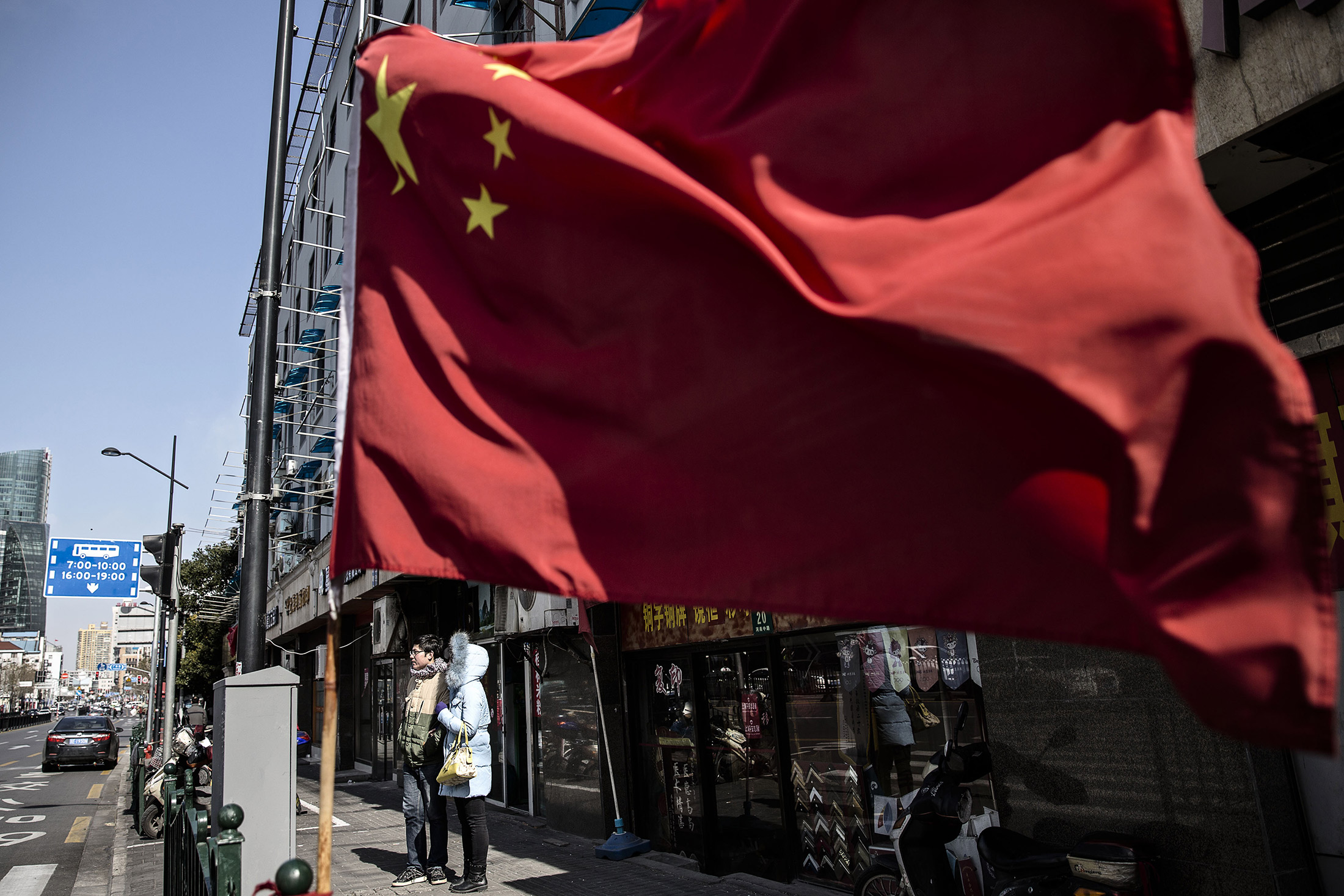 A couple stands on a sidewalk as a Chinese flag flies in Shanghai, China, on Sunday, Jan. 24, 2016. For all the hand wringing over China's economic slowdown, the employment picture has so far remained immune to tumbling stocks, a sliding currency and waning industrial growth.
