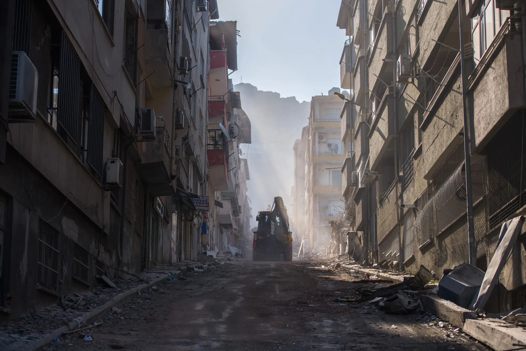 An excavator on&nbsp;a street lined with damaged residential apartment buildings in Hatay, Turkey, in February 2023.