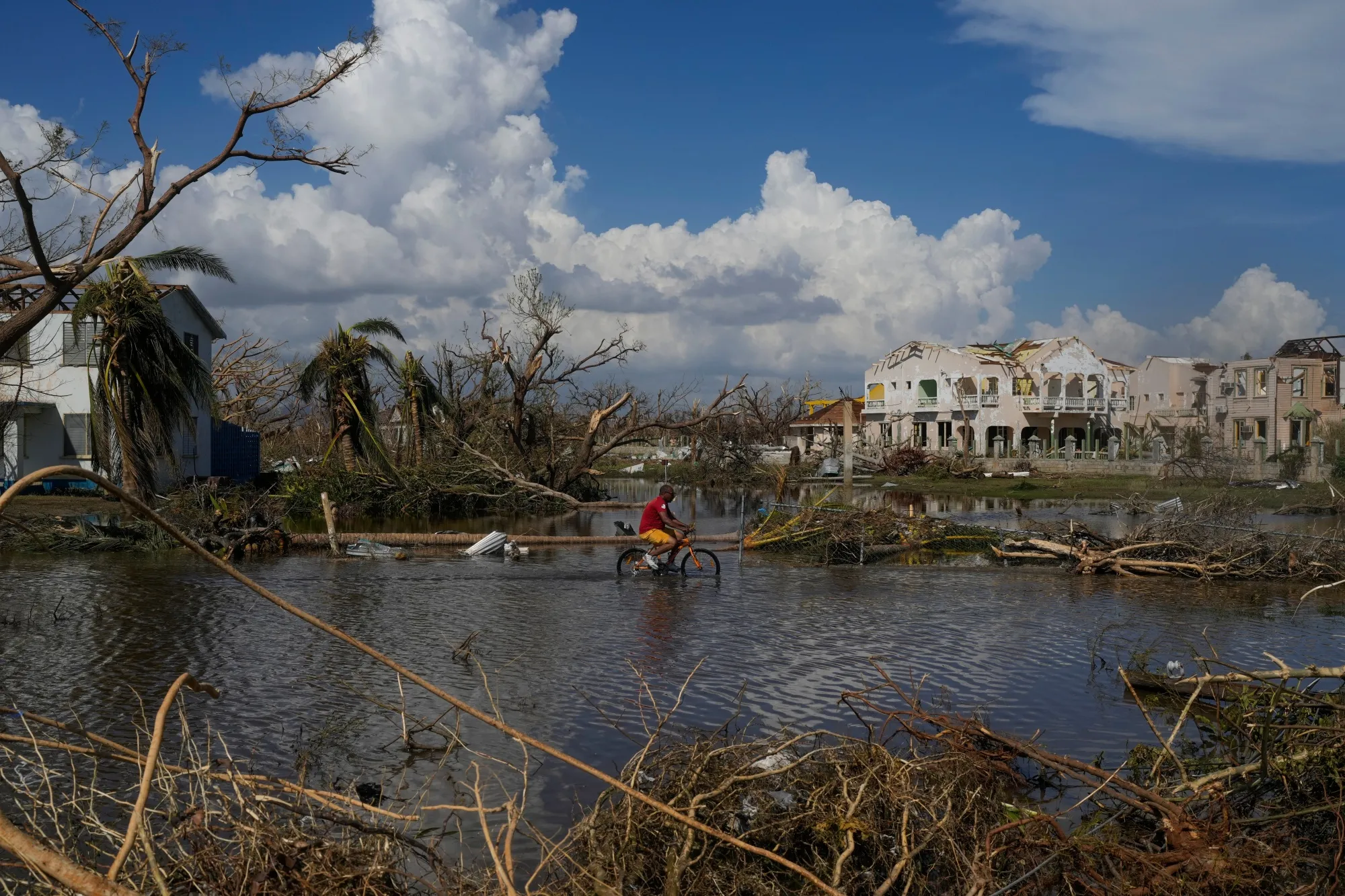 A man is riding a bicycle through a water body under mostly sunny skies with large convective clouds in the background, among buildings without rooves and leafless trees