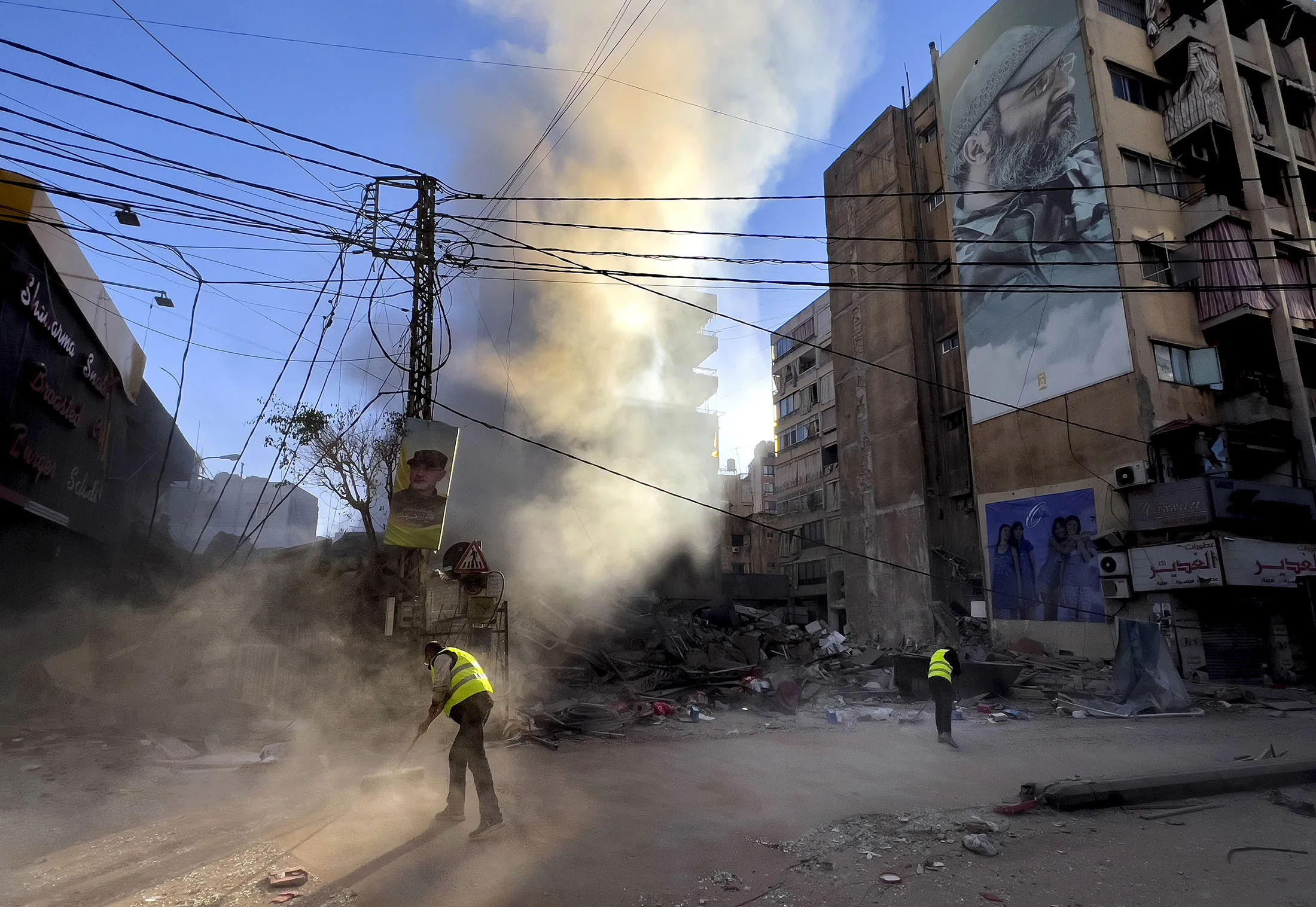 Workers clean a street under a portrait of the late Hezbollah military commander Imad Mughniyeh, as smoke rises from a destroyed building that was hit by an Israeli airstrike in Dahiyeh, the southern suburb of Beirut, Lebanon, on&nbsp;Oct. 20.
