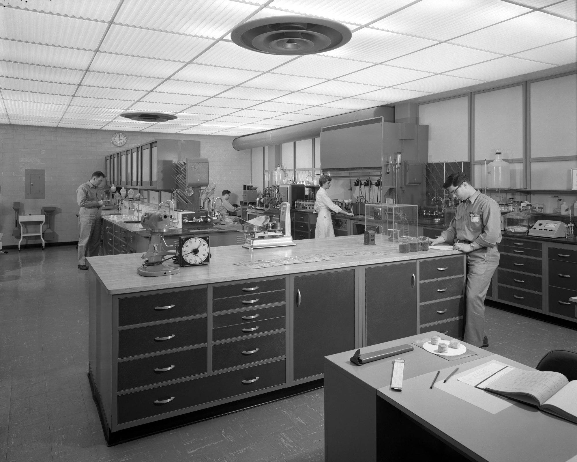People work in a large laboratory with metal cabinets, instruments, and lab benches at Monsanto's St. Louis facility in 1958.