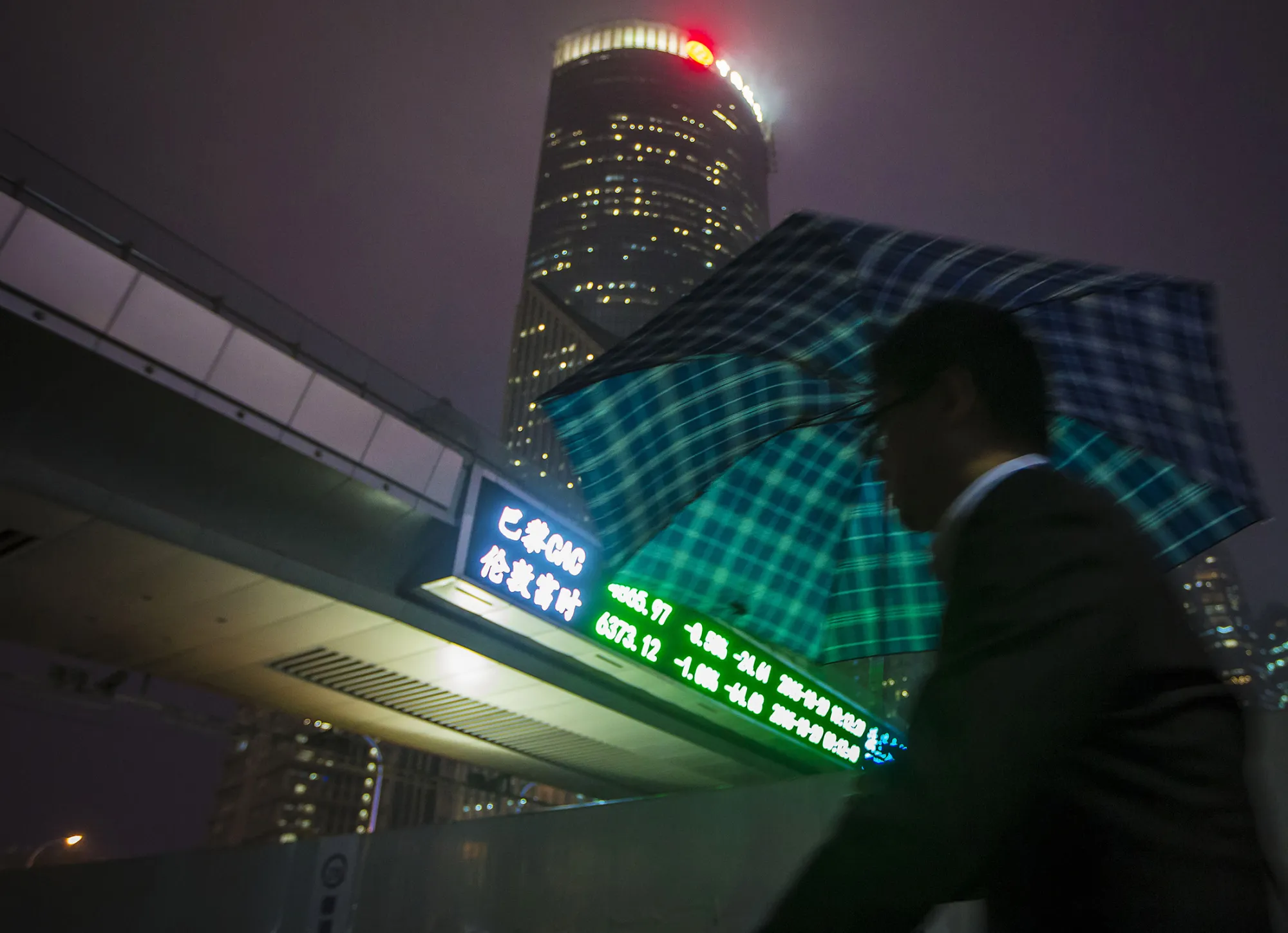 A pedestrian holding an umbrella walks past a pedestrian footbridge featuring an electronic stock ticker at night in the Lujiazui district of Shanghai, China.
