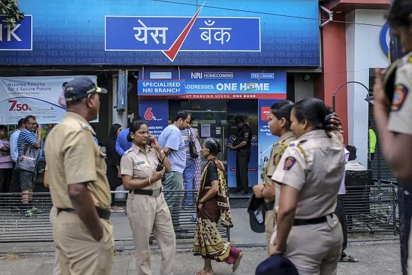 Police officers observe customers standing in line outside a Yes Bank Ltd. branch in Mumbai.