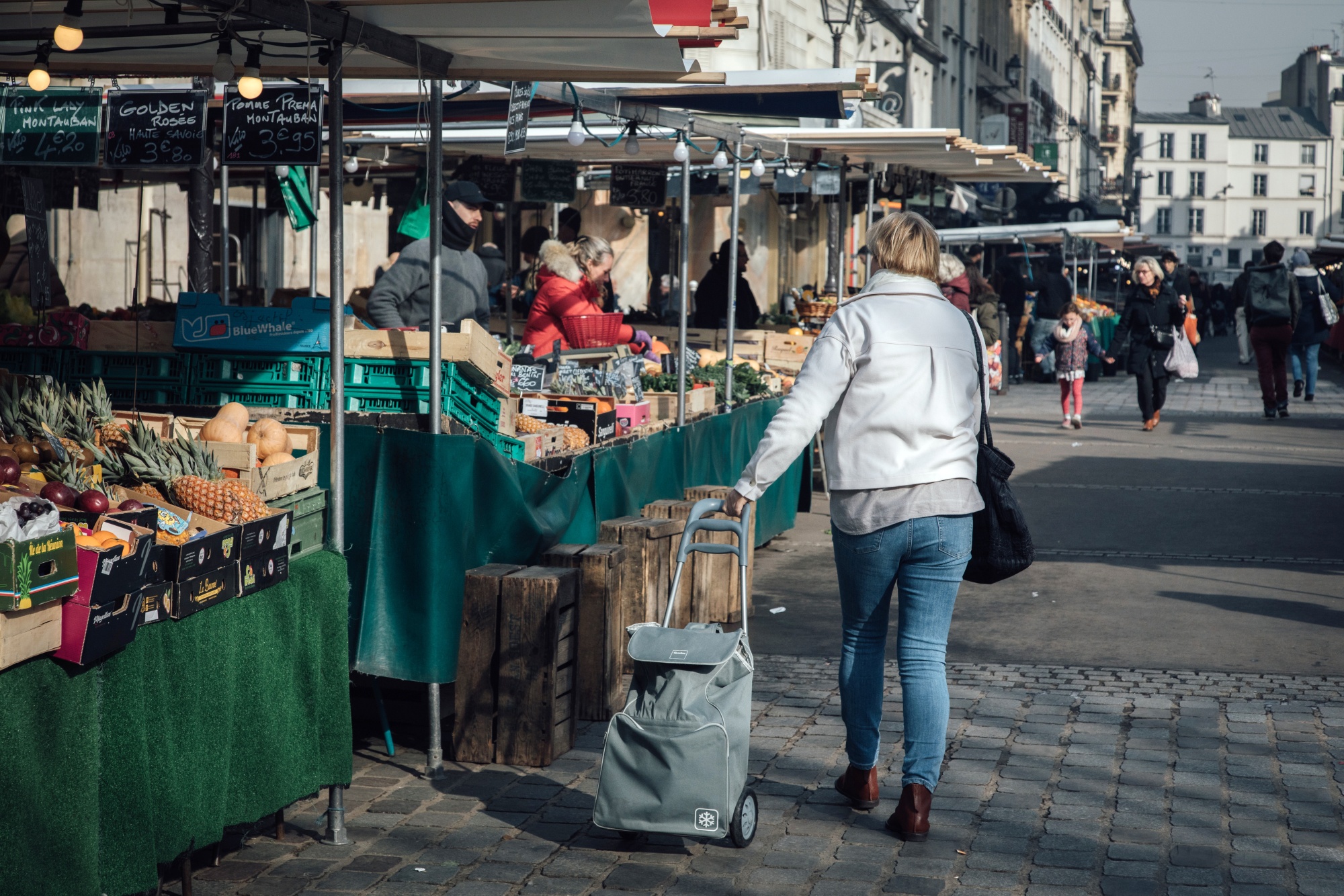 A shopper pulls a trolley bag past past fruit and vegetable stalls in the Bauveau Market in Paris, France.