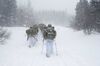 Two columns of soldiers wearing heavy packs snowshoe through a blizzard into the distance, with tall evergreens ahead and large globs of snow being blown into the camera lens.