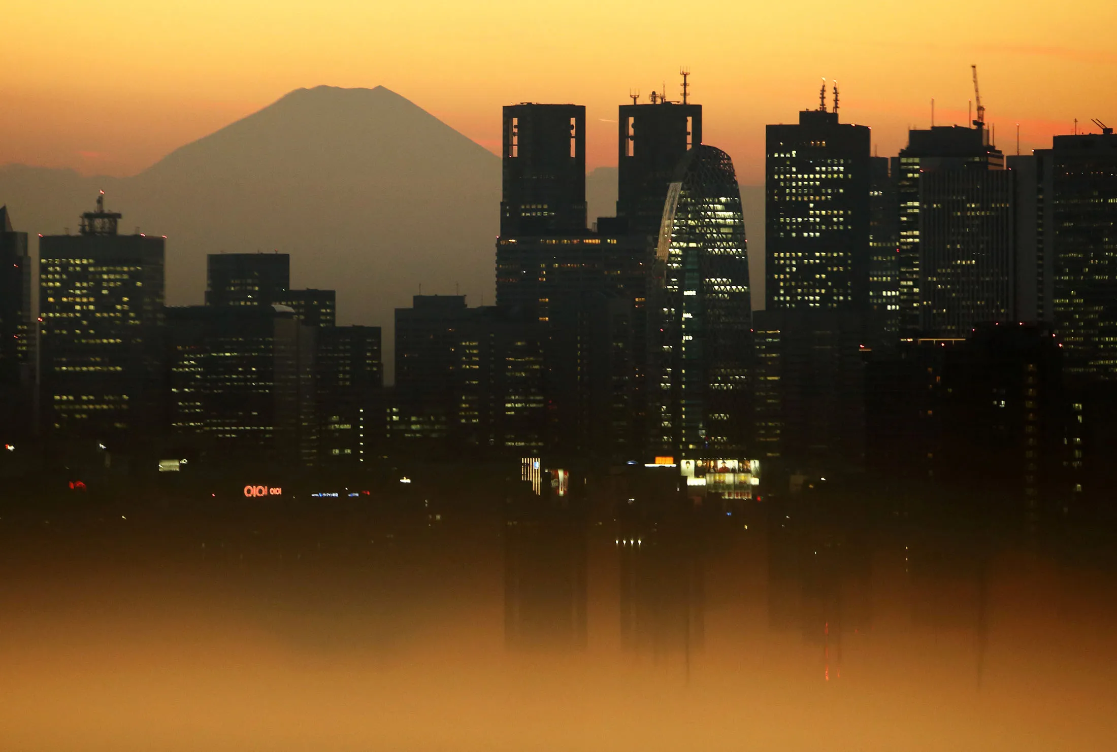 Mount Fuji stands behind illuminated buildings as the sun sets in Tokyo.