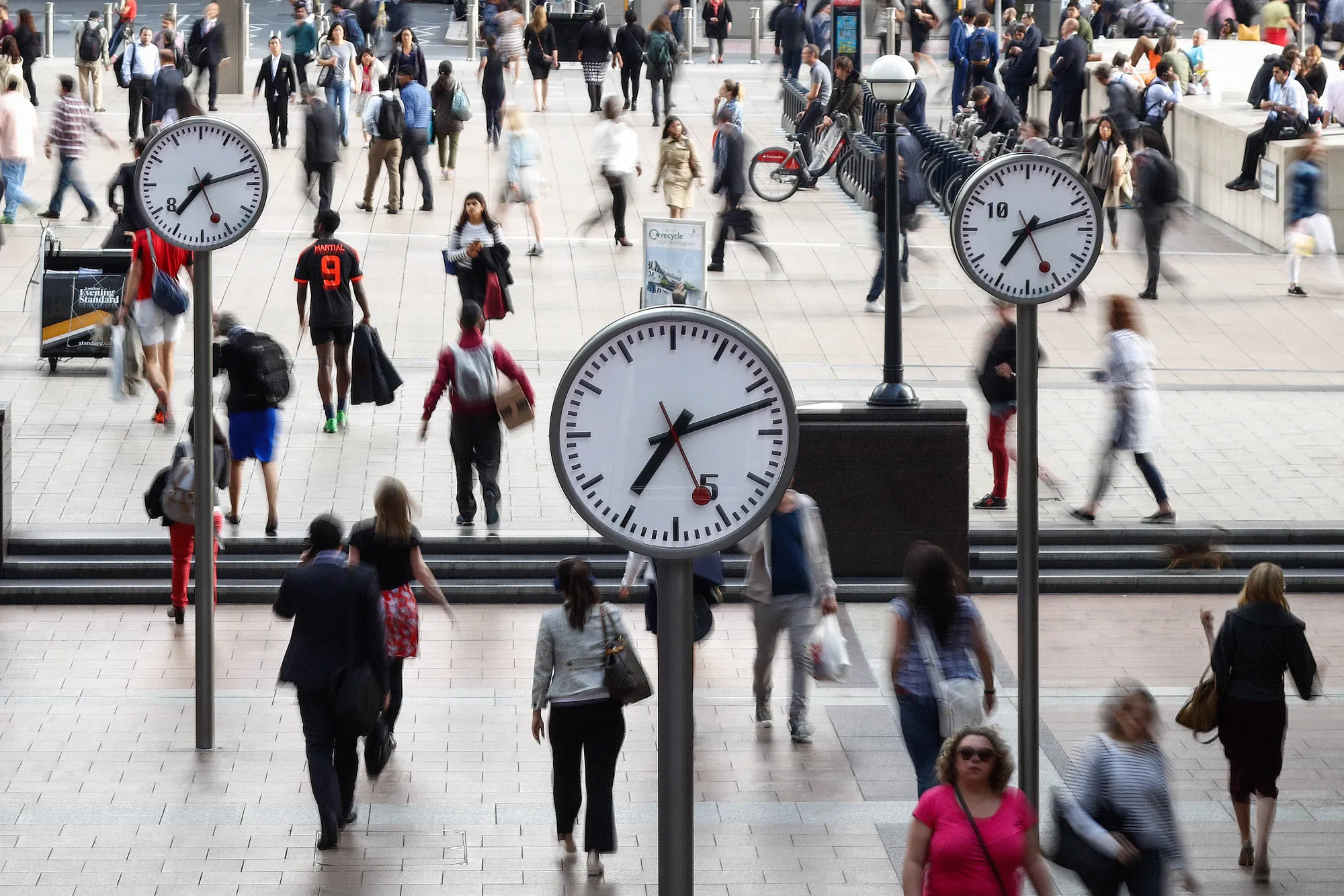 Pedestrians walk past clocks in the Canary Wharf financial, shopping and business district in London, U.K.