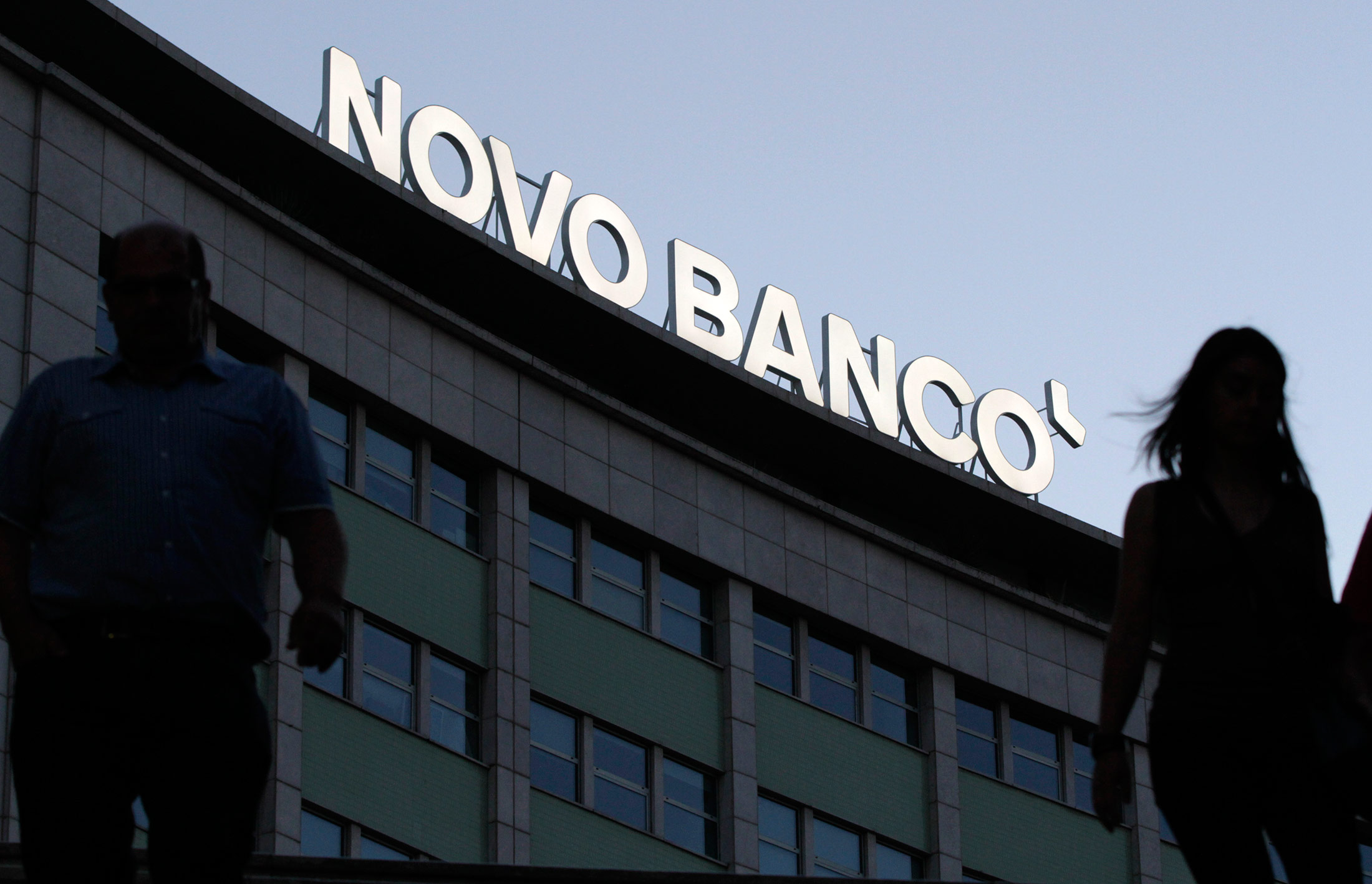 Pedestrians pass an illuminated Novo Banco SA logo above a building in Lisbon, Portugal