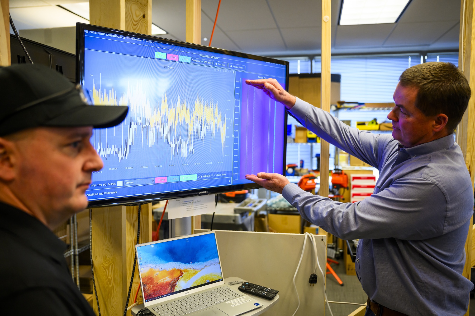 Whisker CEO Marshall is a caucasian male with trim hair, wearing a blue-gray button-down shirt and standing to the right of a large monitor with his hands spread apart vertically measuring the distance between two points on a chart that is shown on the screen, representing real-time voltage data. Under the monitor is a laptop and some remote controls, and in the foreground is another caucasian male wearing a black baseball cap with some reading glasses resting above the brim and wearing a black polo shirt. Both men are visible in a profile view, looking in opposite directions.