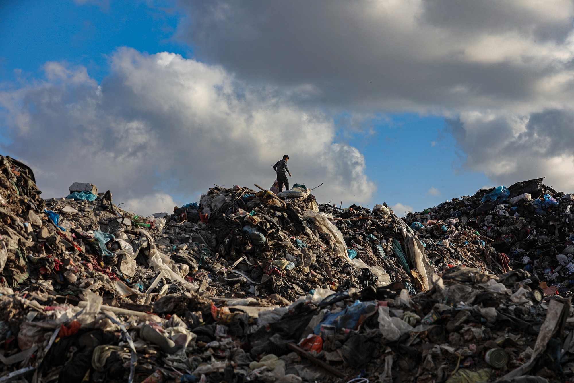 A photograph of a person walking across the top of a sprawling trash site. The lone figure is small and silhouetted against a dramatic, cloudy sky. The image shows a person searching for salvageable items at the site of the former Souk Feras market in Gaza City dated April 2025.
