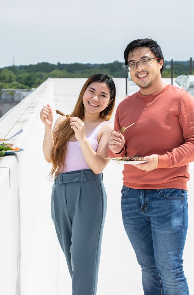 Sokhhita Sok, wearing pink tank top and light blue pants, with friend wearing salmon shirt and blue denim. Posing for picture on roof holding beef skewers