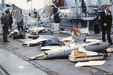 Salvaged debris from Air India Flight 182 arrayed on a dock outside a navy vessel with various officials surrounding the debris.