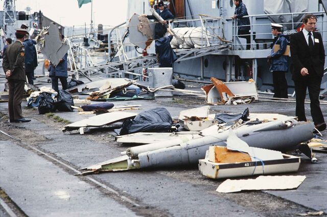 Salvaged debris from Air India Flight 182 arrayed on a dock outside a navy vessel with various officials surrounding the debris.