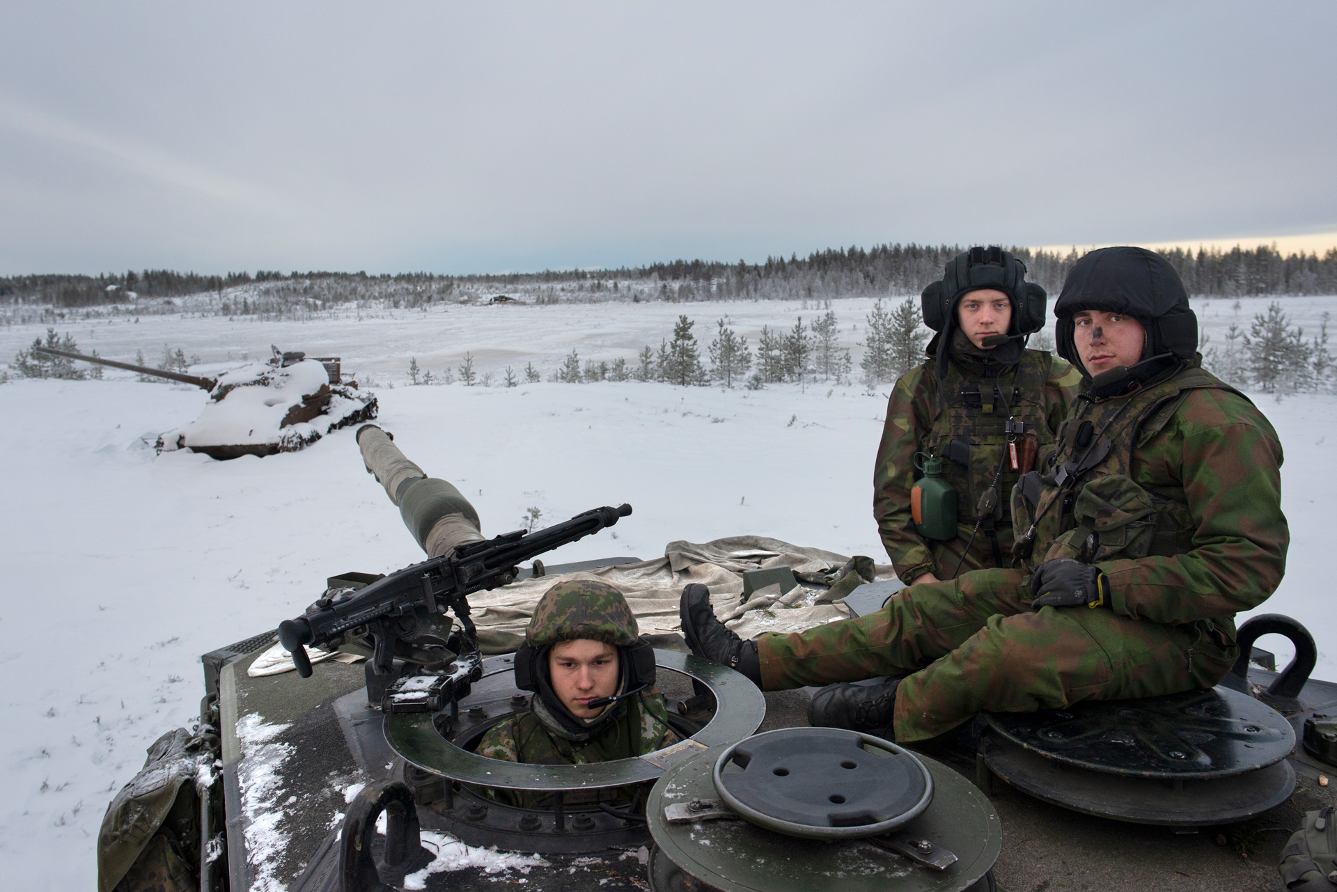 Three Finnish soldiers, facing the camera and wearing green camouflage uniforms, helmets and radio headsets are on top of a tank in a snow-covered wilderness. One pokes his head up through an open hatch while the other two sit up top to one side. The muzzle of the tank's gun points toward an old tank half-buried in snow a short distance away.