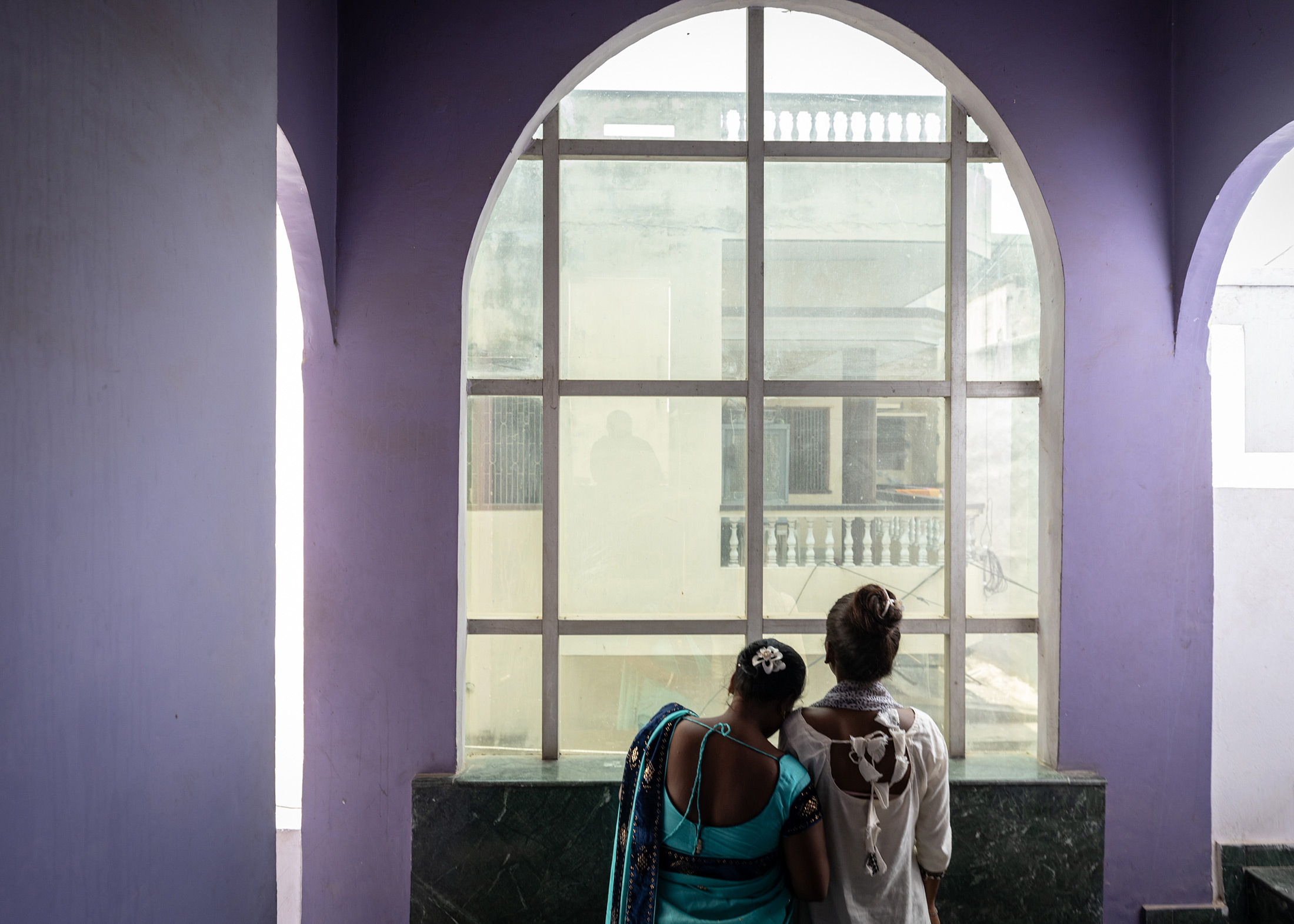 An older woman in a blue green sari rests her head on the shoulder of a younger woman dressed in white. Both have their back to camera and are looking out a window. 