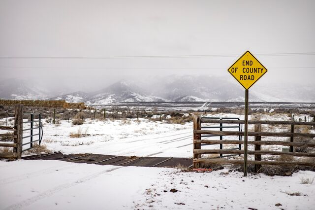 A photo of a road that reaches and passes through a fence by way of an open gate. There is a sign at the fence that says "End of county road." The ground is snow-covered, as are mountains in the distance.