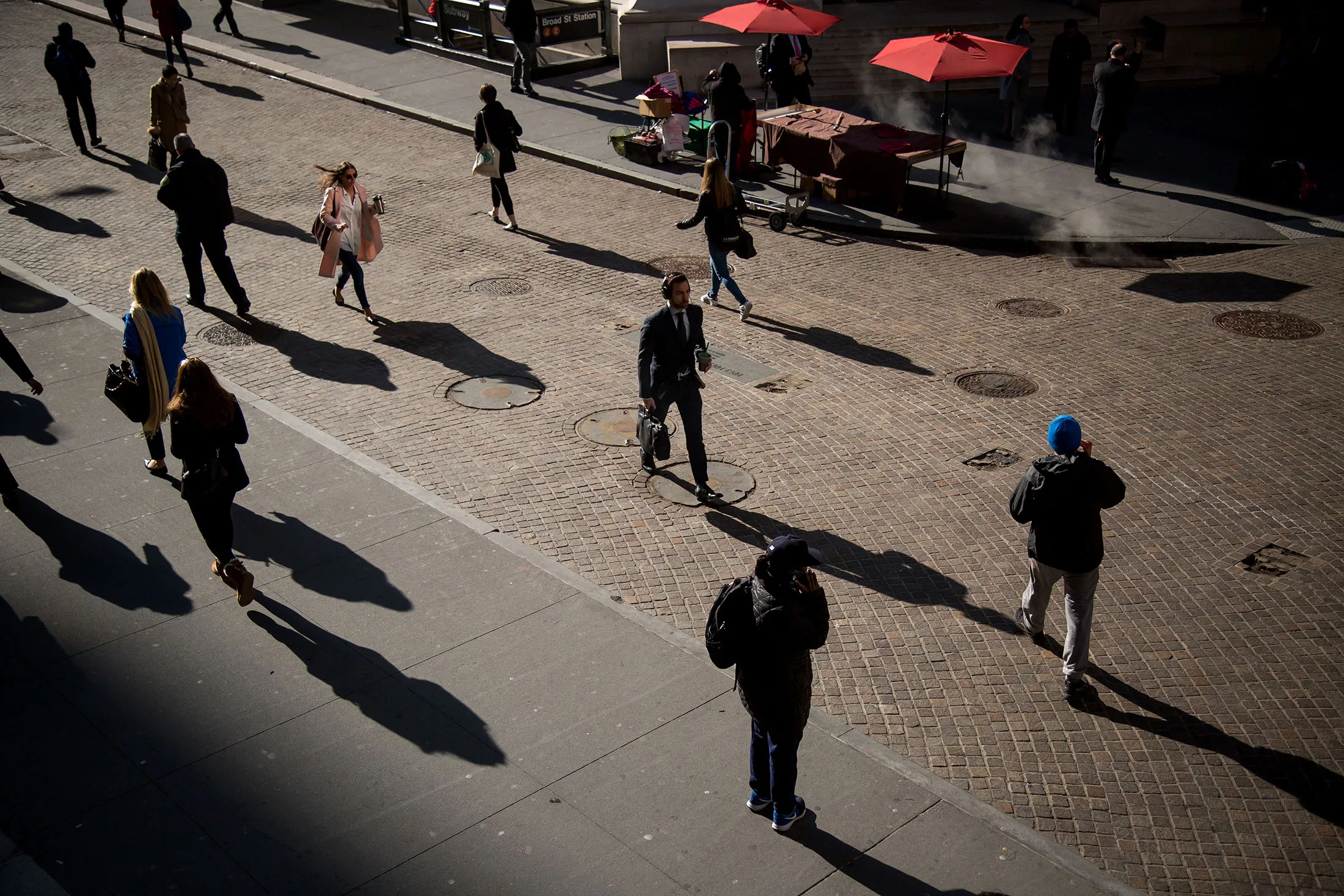 Pedestrians cast shadows as they walk on Wall Street near the New York Stock Exchange (NYSE) in New York, U.S., on Monday, March 7, 2016. U.S. stocks fluctuated near two-month highs as commodity shares surged with crude oil, offsetting declines among technology and consumer companies as investors assessed China's growth prospects.

