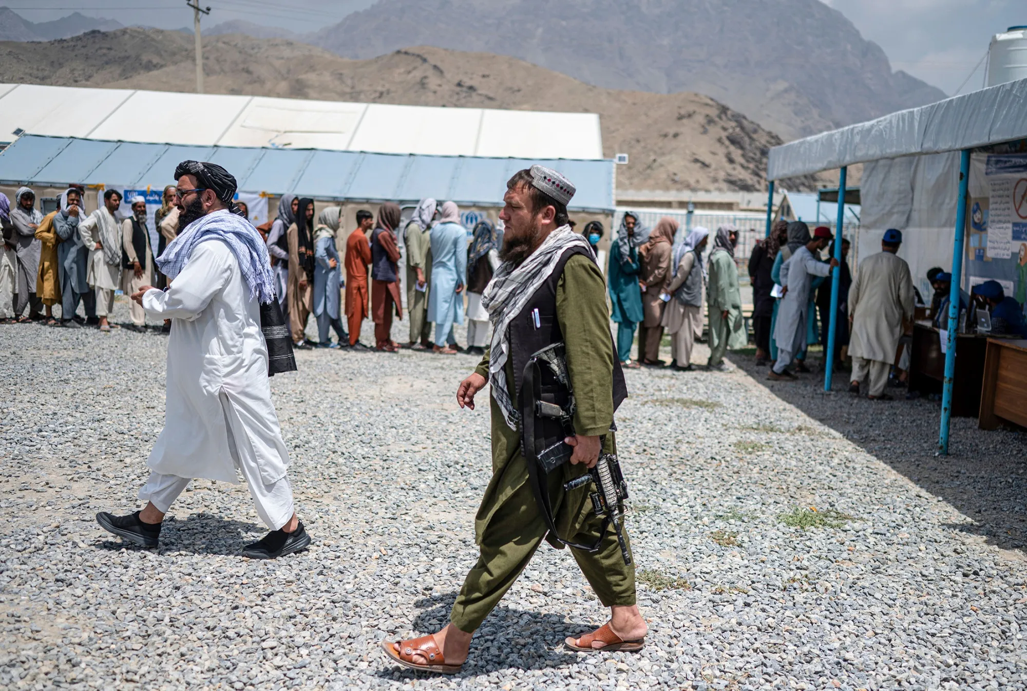Taliban fighters walk past internally displaced refugees in a queue at the UNHCR camp in the outskirts of Kabul on July 28.