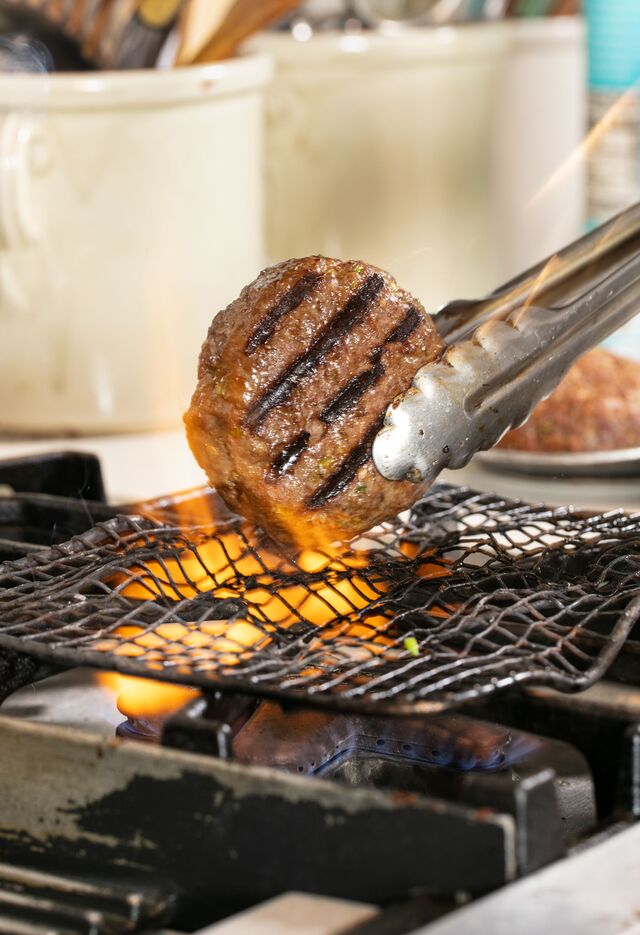 Image shows a hamburger being cooked over an open flame grill.