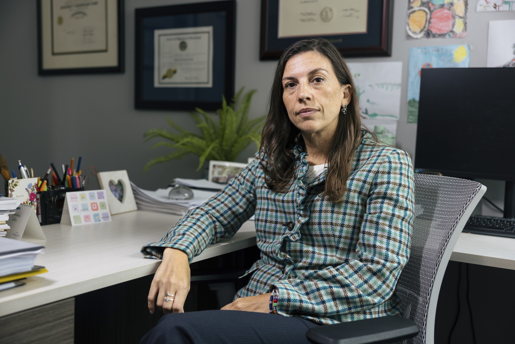 A woman with brown hair, wearing a checkered suit, sits at her desk and rests her arm on the desk, behind her on the wall are framed images of her certificates and on the desk there are cards and stacks of paperwork.