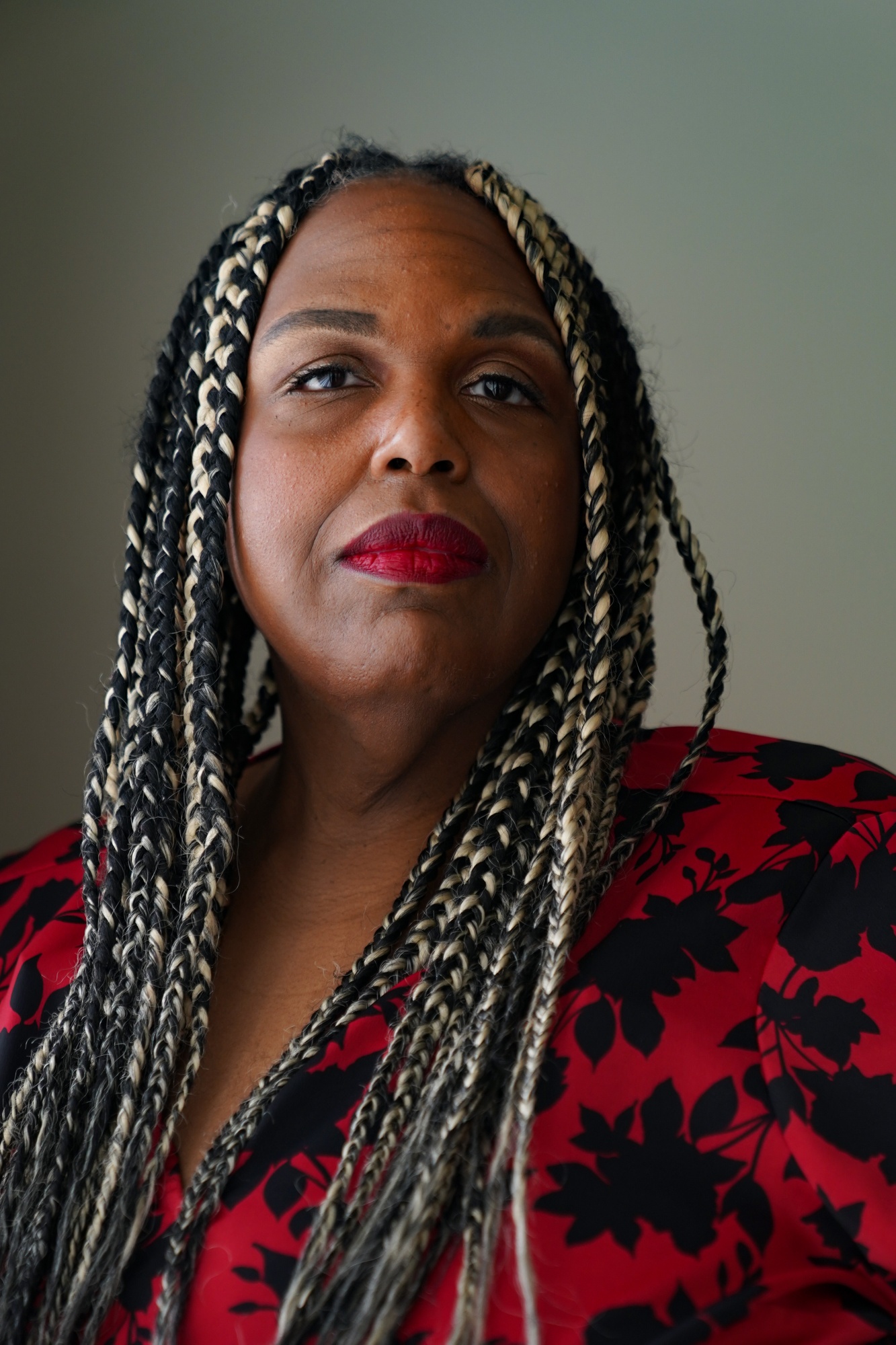 A portrait of a black woman with locked black and golden hair, wearing a red outfit with black flower print, and red lipstick. She is indoors and appears to be next to a window that is out of frame