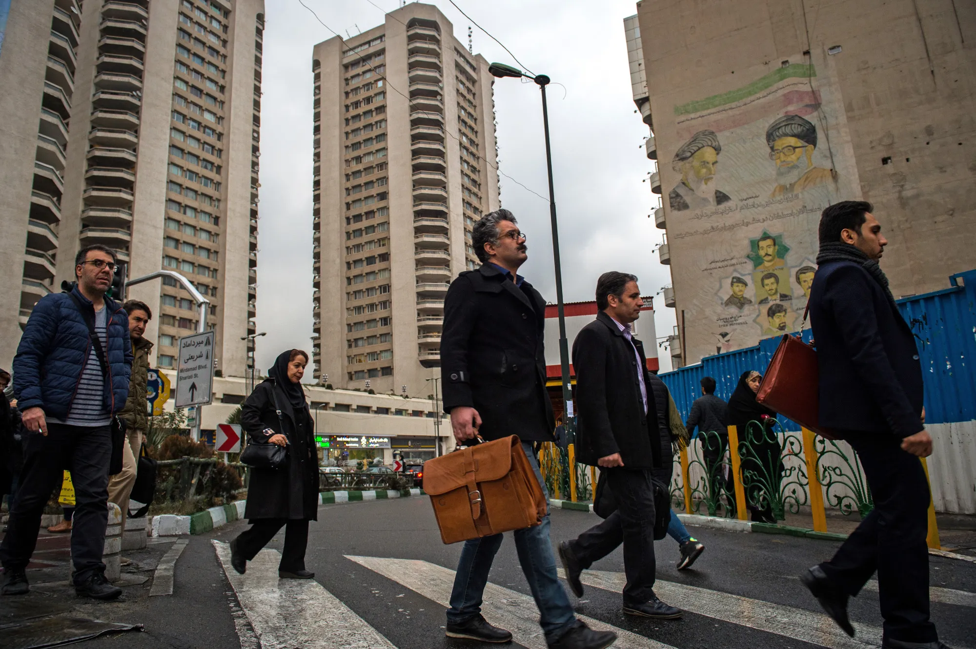 Commuters pass under neath tower blocks and faded banners depicting Iran's religious leaders while crossing a street in Tehran.