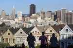Pedestrians stand in front of Victorian homes and the downtown skyline in San Francisco.