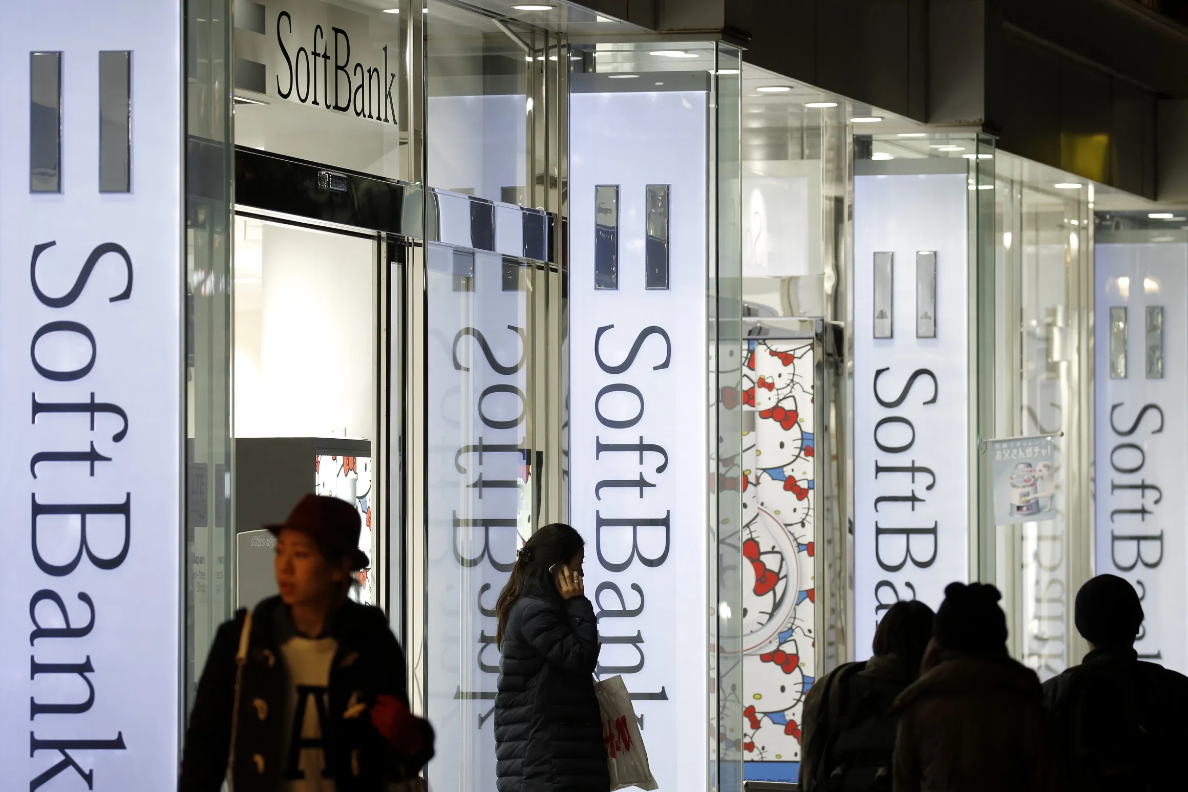 A customer, center, uses her smartphone as she exits a SoftBank Corp. store at night in Tokyo, Japan, on Sunday, Feb. 8, 2015. SoftBank, the Japanese wireless carrier led by billionaire Masayoshi Son, is scheduled to report third-quarter earnings on Feb. 10.
