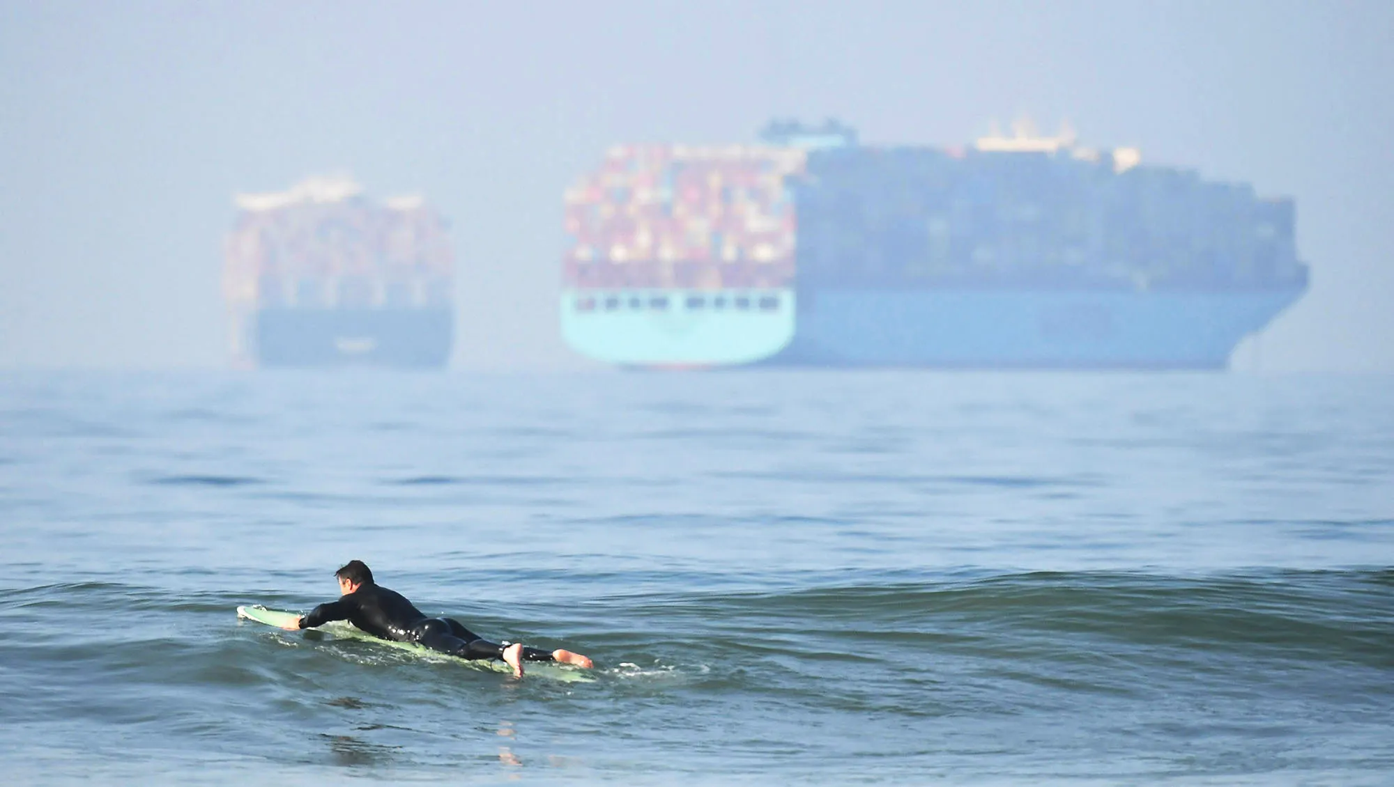 A surfer waits for waves at Huntington Beach as container ships wait offshore.