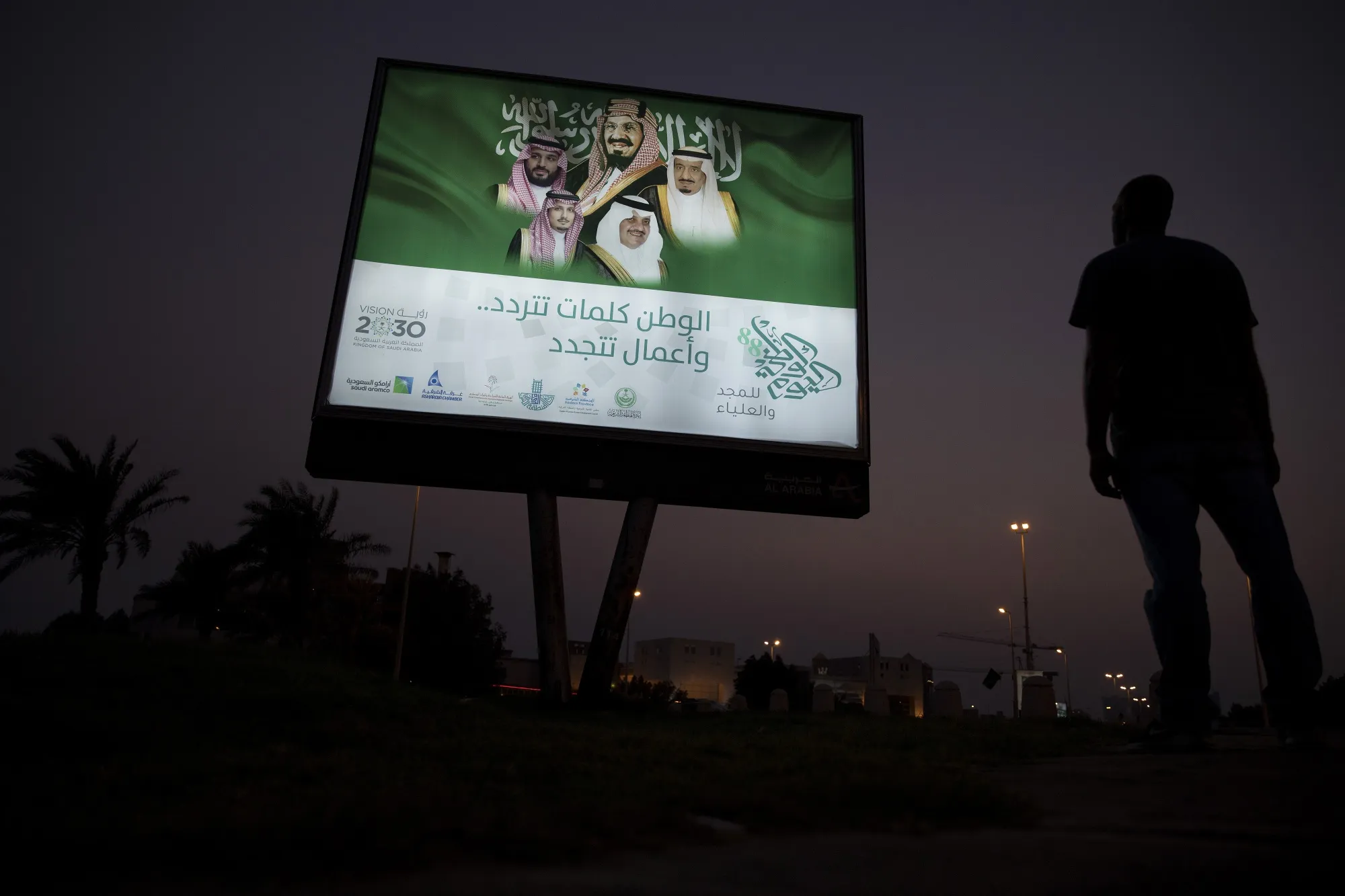 A pedestrian pauses near an illuminated sign showing portraits of the Saudi Arabian Royal family in Dhahran, Saudi Arabia.