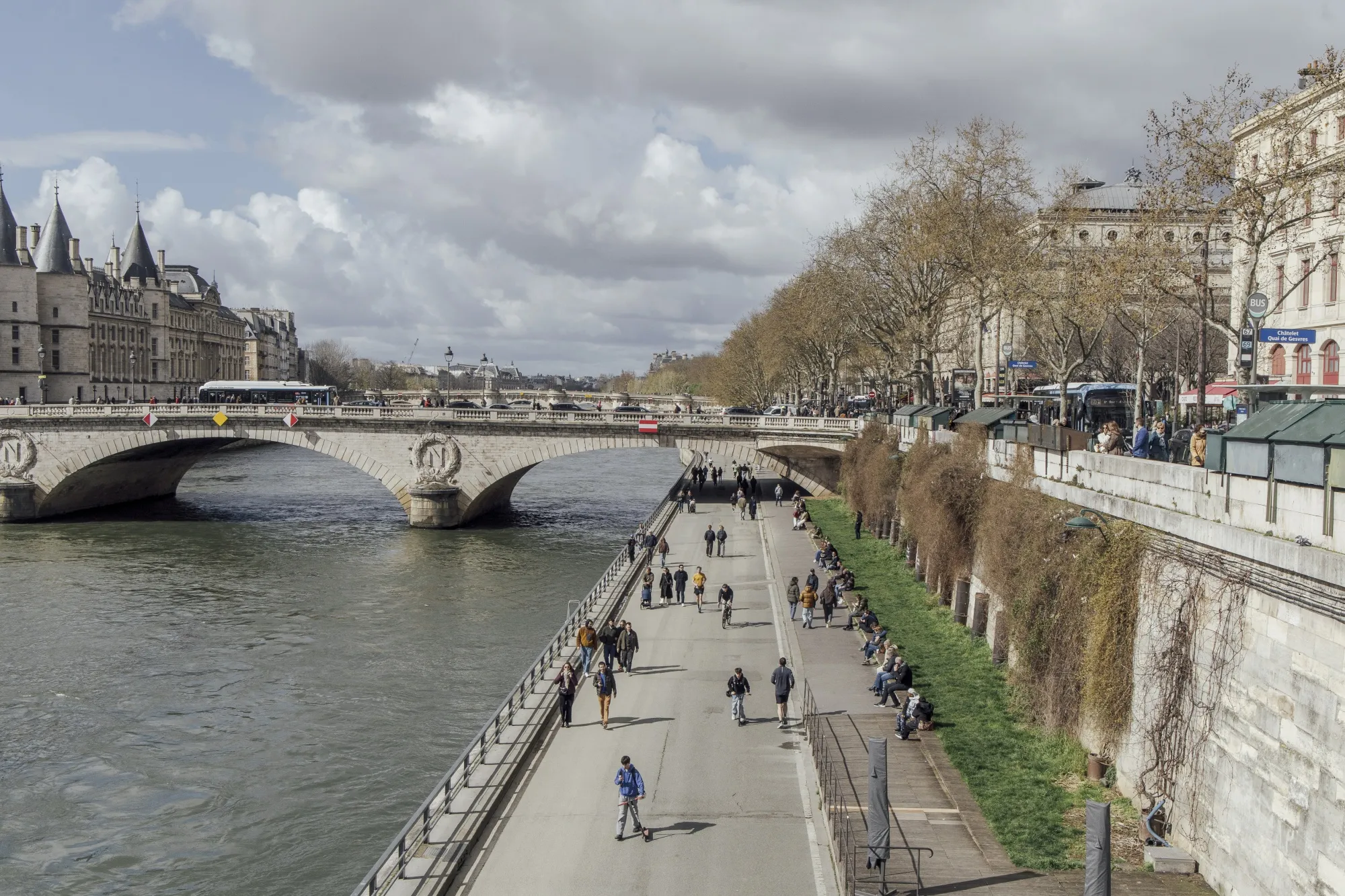 A high-angle view of the Seine River in Paris on a bright, partly cloudy day in March. To the left, a stone arch bridge carrying a bus spans the water toward the turrets of the Conciergerie building. On the right, a wide pedestrian promenade runs parallel to the river, bustling with people walking and lounging on a grassy embankment. Bare spring trees and traditional Parisian building facades line the upper quay.