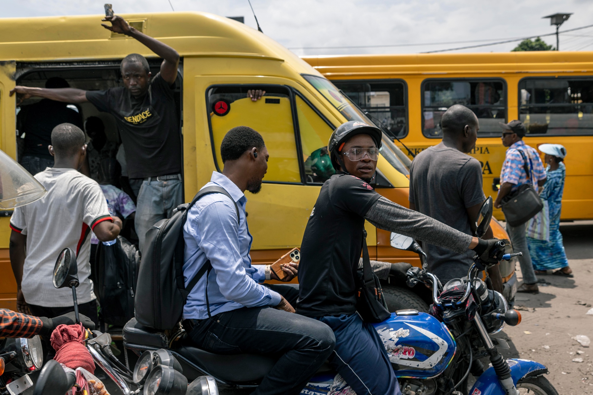 Motorcycle taxi riders waiting for passengers near yellow buses in Kinshasa.