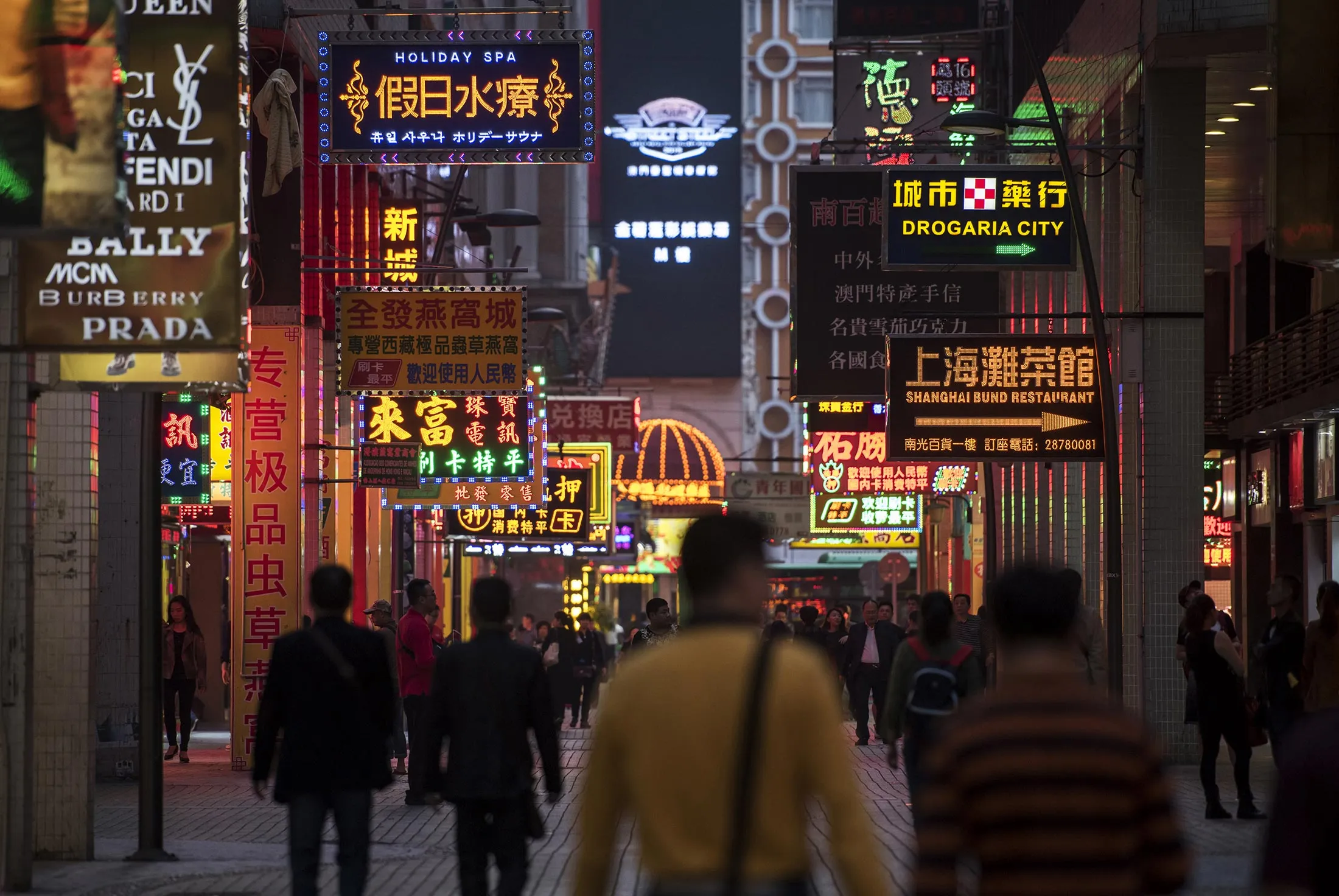 Pedestrians walk along a street with illuminated signs at night in Macau, China, on Tuesday, Dec. 1, 2015. Macau's casino revenue fell for the 18th straight month in November, as China's moves to curb illicit money flows from the mainland deterred the high-stakes players who rely on junket promoters for betting loans.
