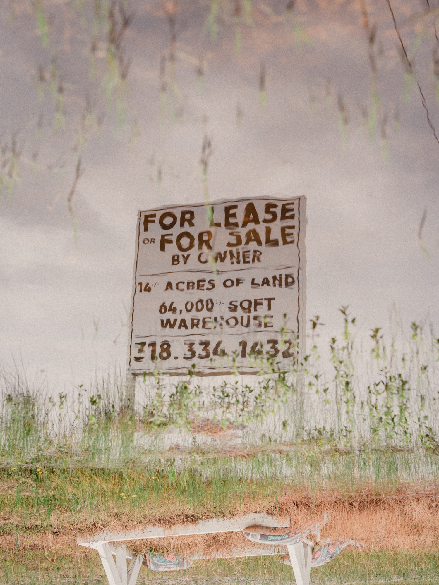 A blurry reflection in water of a “For Sale” sign advertising thousands of acres for sale.