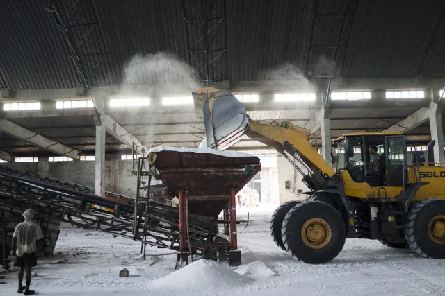 A wheel loader moves urea fertilizer onto a conveyor inside a warehouse at a port in India.
