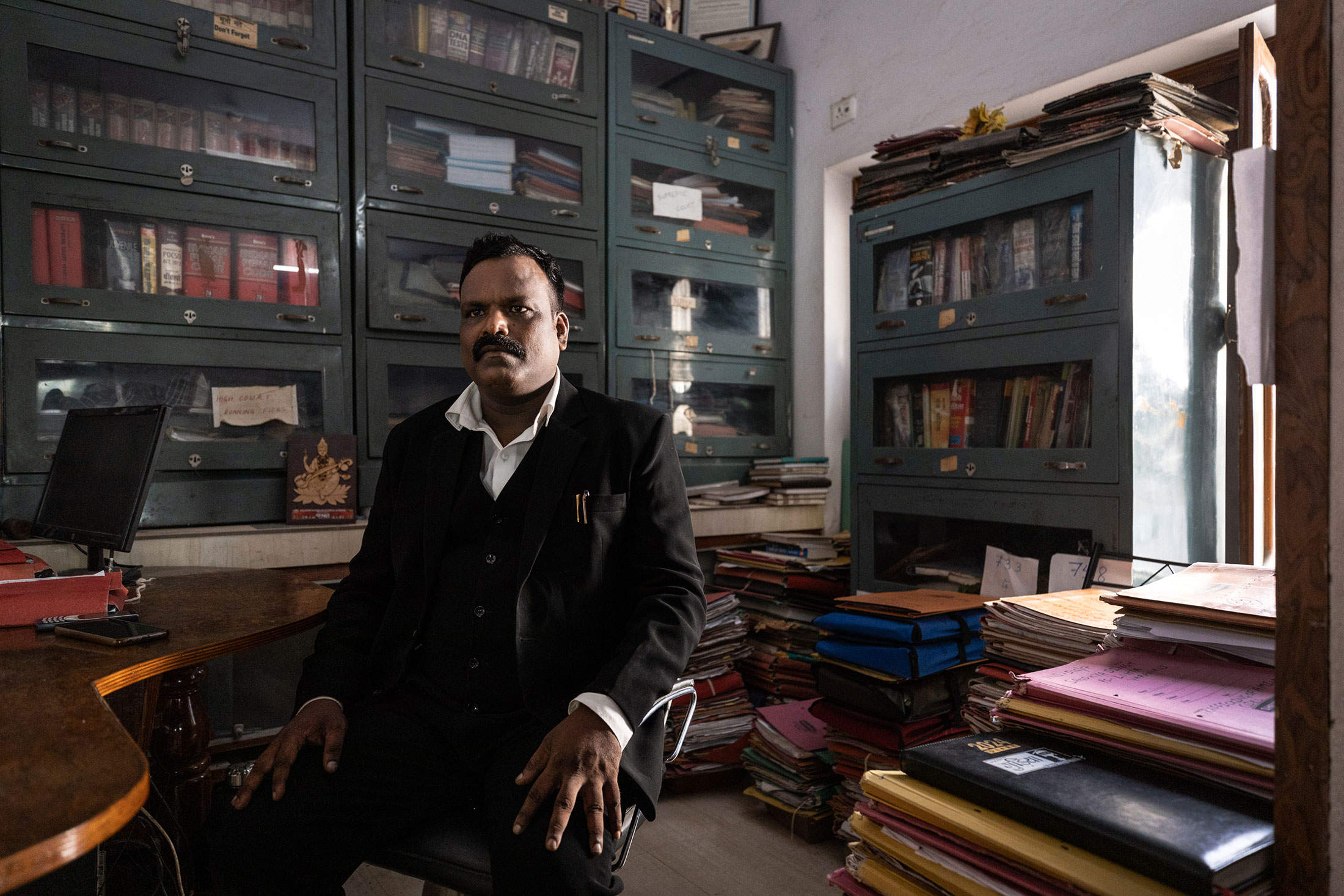 A man with dark skin and a moustache wearing a black suit and white shirt with two pens in the pocket sits on a chair with his hands on his knees. He is facing the camera and is surrounded by stacks of folder files of many colors.